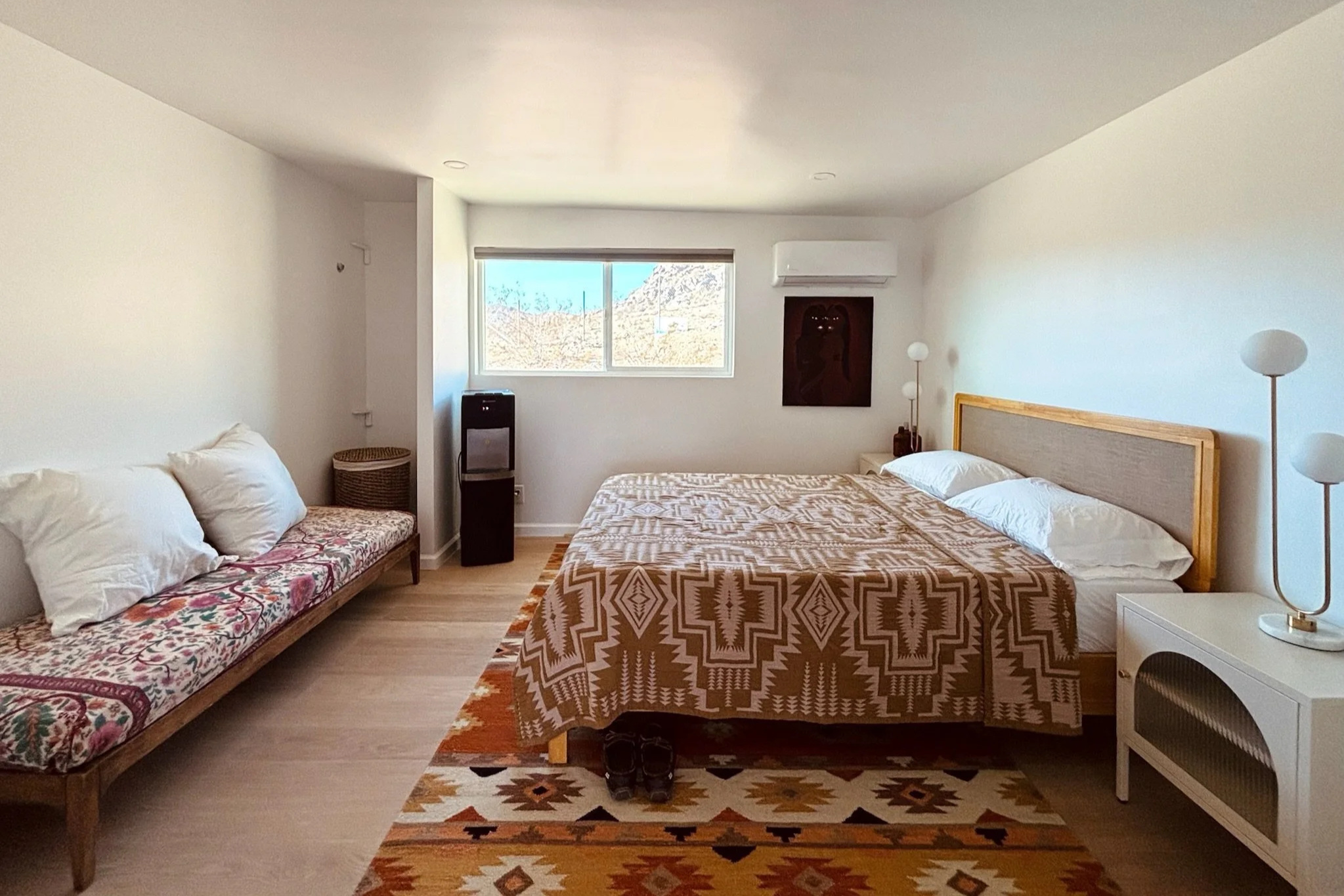 A bright minimalist bedroom with a bed, nightstand, lamps, window, and a bench with pillows.