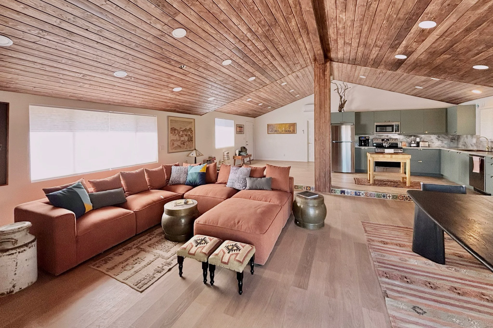 Living room with orange sectional sofa, throw pillows, a wooden coffee table, and a rug, opening to an kitchen with green cabinets, stainless appliances, and a small wooden table, all under a wood-paneled ceiling.