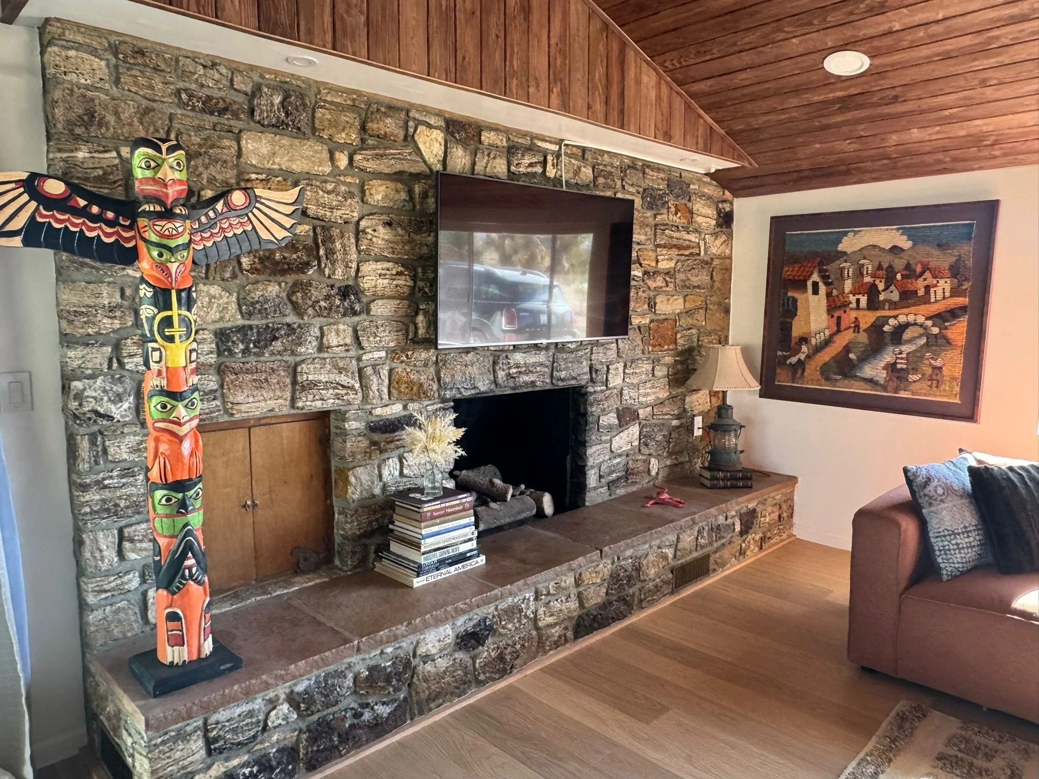 Living room with a stone fireplace, a mounted flat-screen TV, a colorful Native American style totem pole, a stack of books with an arrangement of dried flowers, a table lamp, a framed artwork of a village scene, a beige sofa with pillows, and wooden