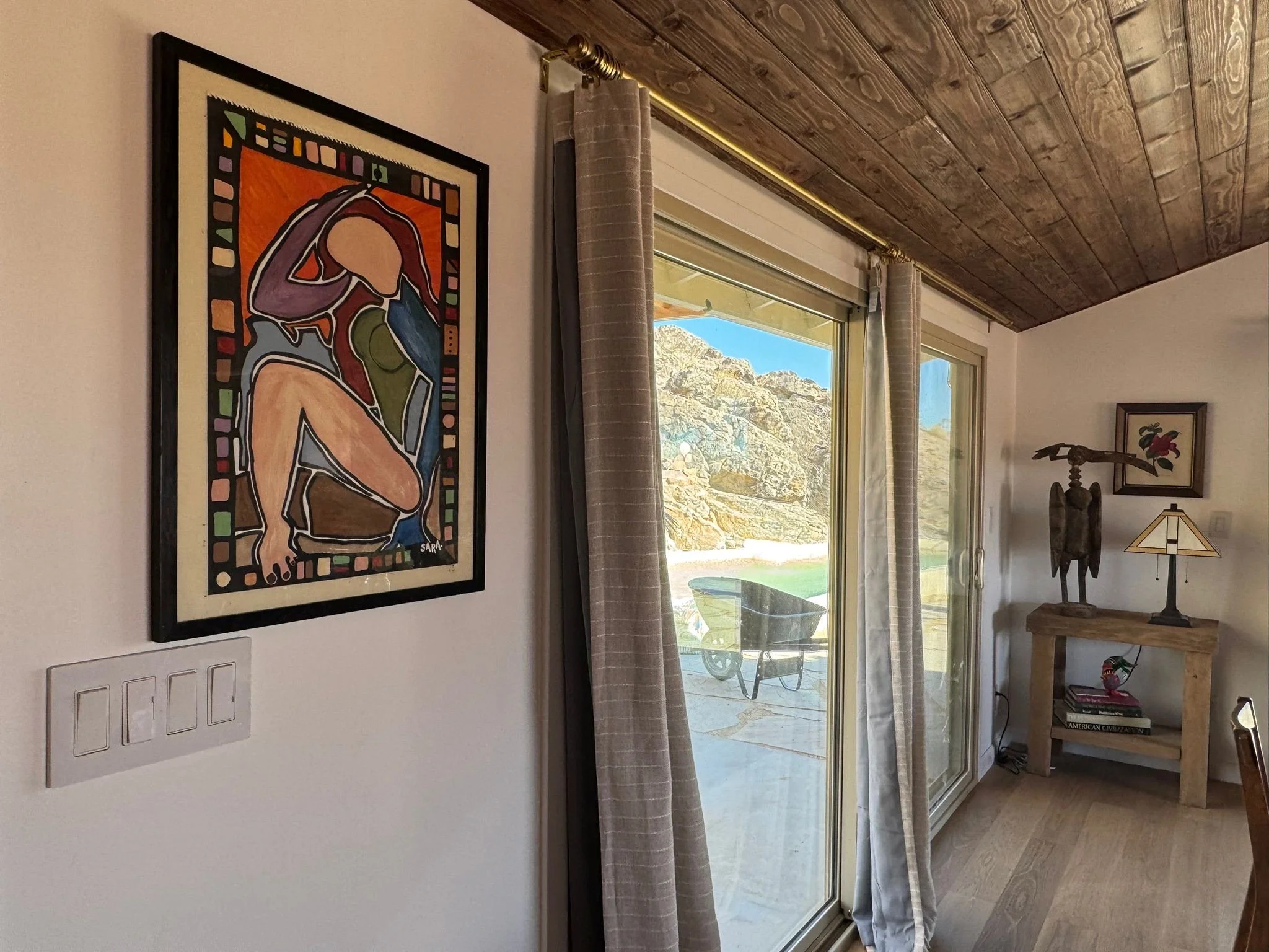 Interior of a room with a sliding glass door showing an outdoor patio with rocky landscape and water. Inside, there is a colorful abstract art piece on the wall, beige curtains, a wooden console table with sculptures, books, and a lamp.