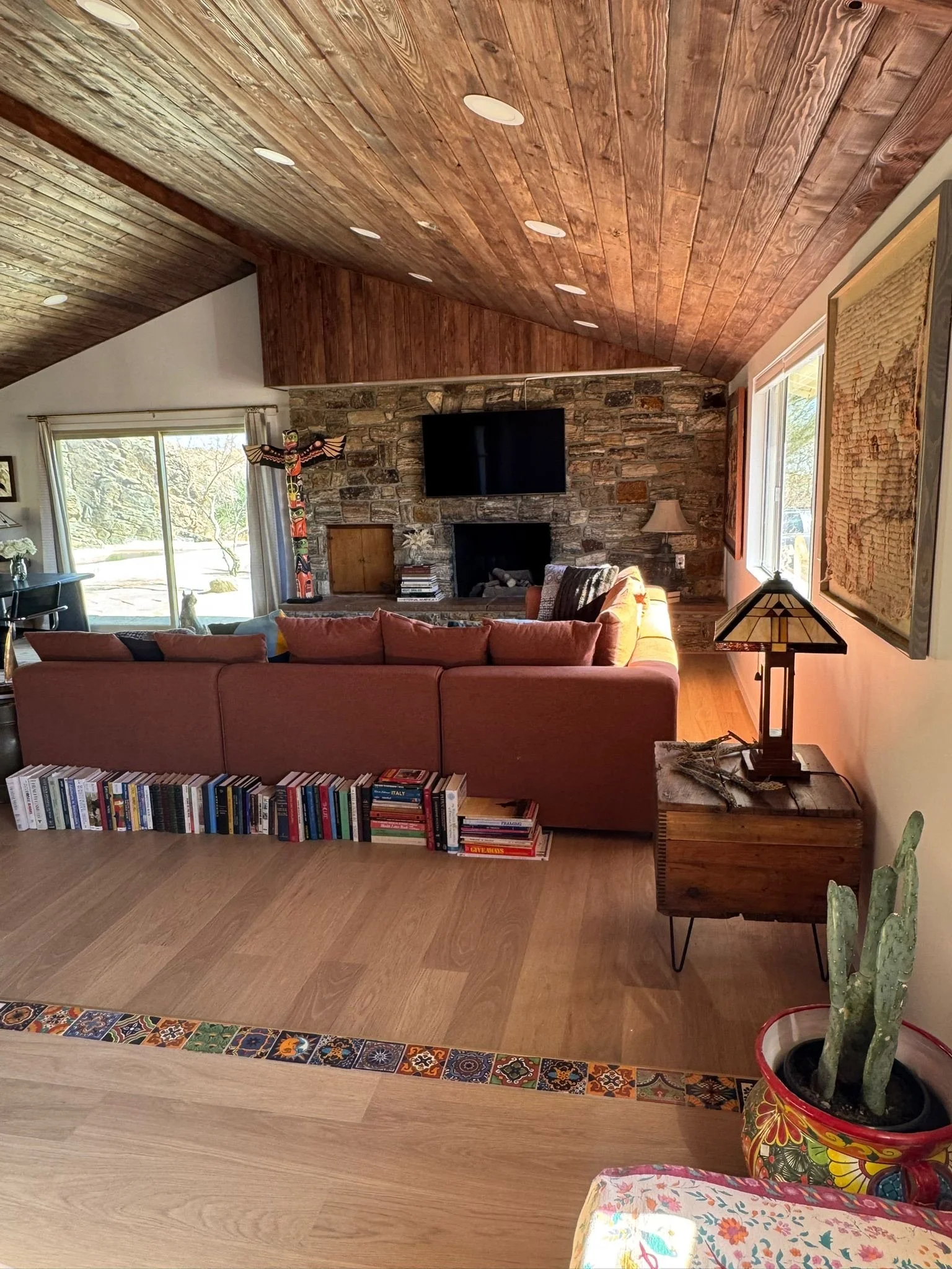 Living room with wooden ceiling, stone fireplace, television, sofa, multicolored bookshelf, side table with lamp, large window, potted cactus, colorful rug.