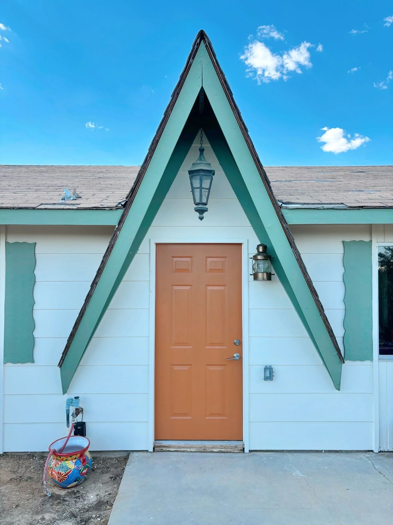 Front view of a house with a unique A-frame roof, brown door, hanging lantern light, and an outdoor lantern on the right side. A colorful bucket with gardening tools is on the ground left of the door.