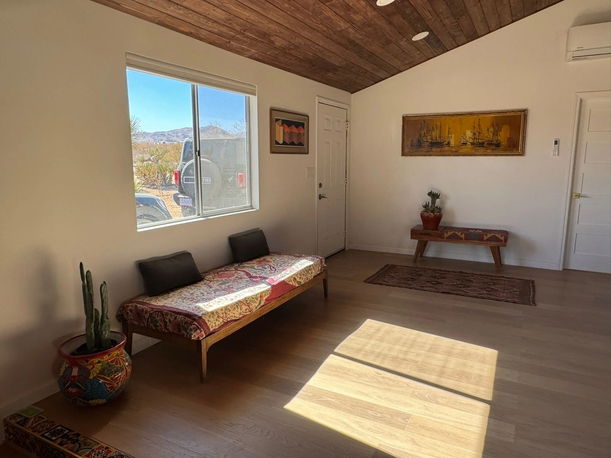 Sunny interior living room with wooden ceiling, white walls, and light wood flooring. A window showing a desert landscape with mountains and two parked vehicles, including a Ford truck. Decor includes a cactus in a colorful pot, a bench with a patter
