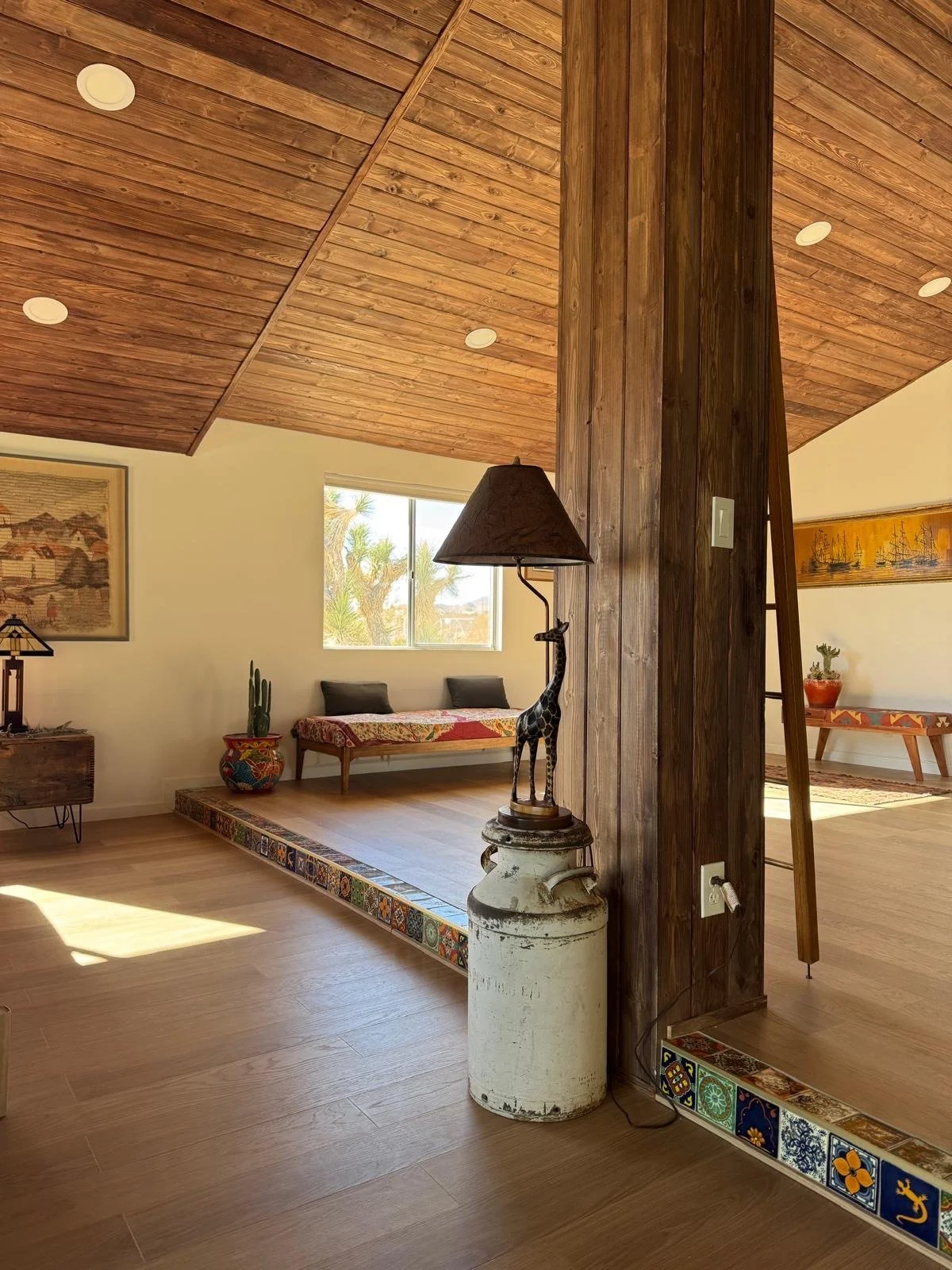 A cozy living room with wooden ceiling and floor, a window, and a white wall. There is a wooden bench with pillows, a cactus in a decorative pot, and a giraffe-shaped lamp on an old milk can. Colorful patterned tiles line the doorway, and artwork and