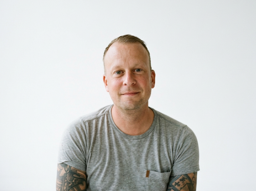 A man with short hair wearing a gray t-shirt, standing against a plain white background, looking at the camera with a slight smile.