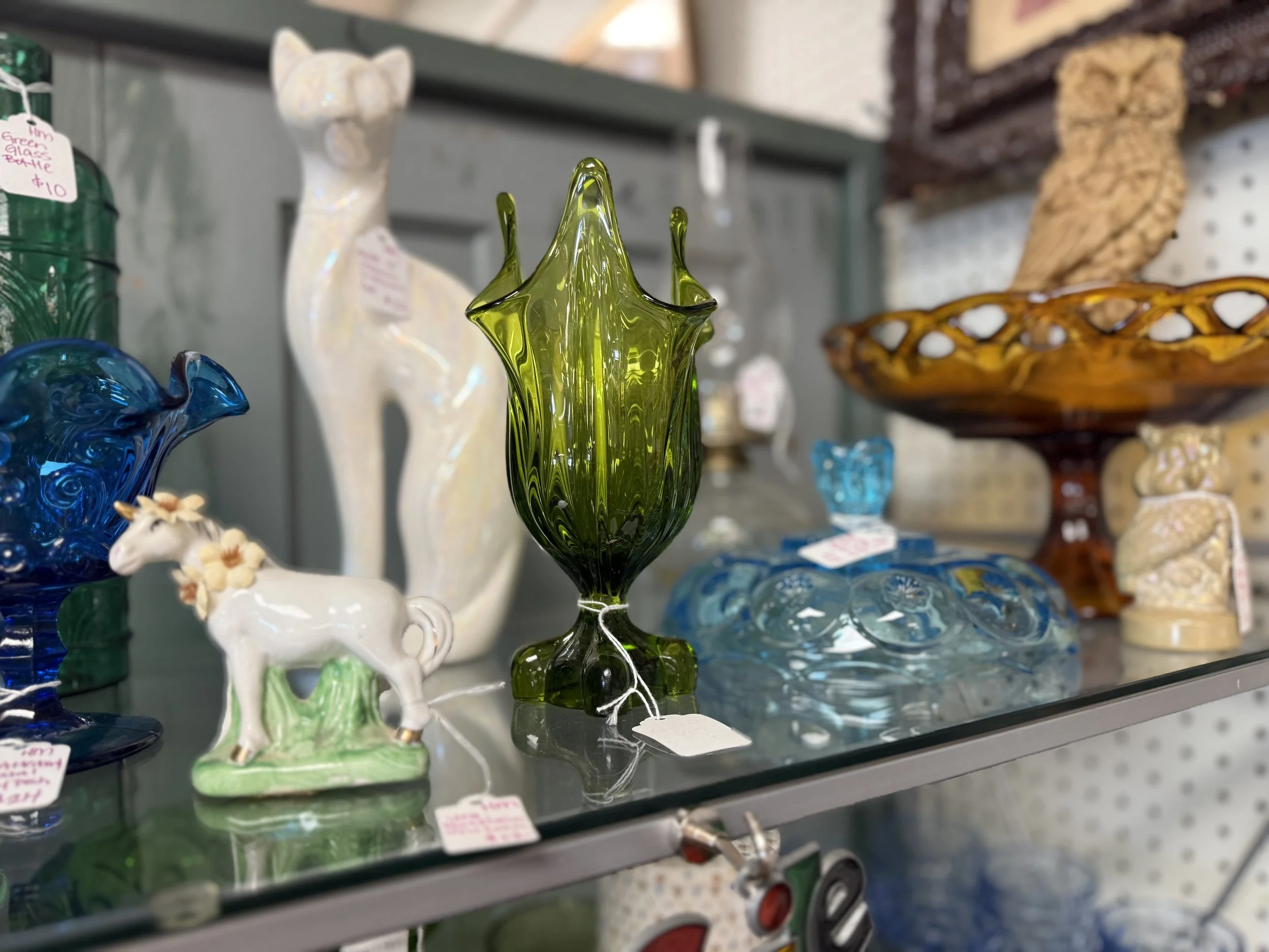 Colorful glass vases and figurines displayed on a glass shelf in a store. A prominent green glass vase with a floral shape is in the center, with smaller blue, white, and brown glass objects around it.