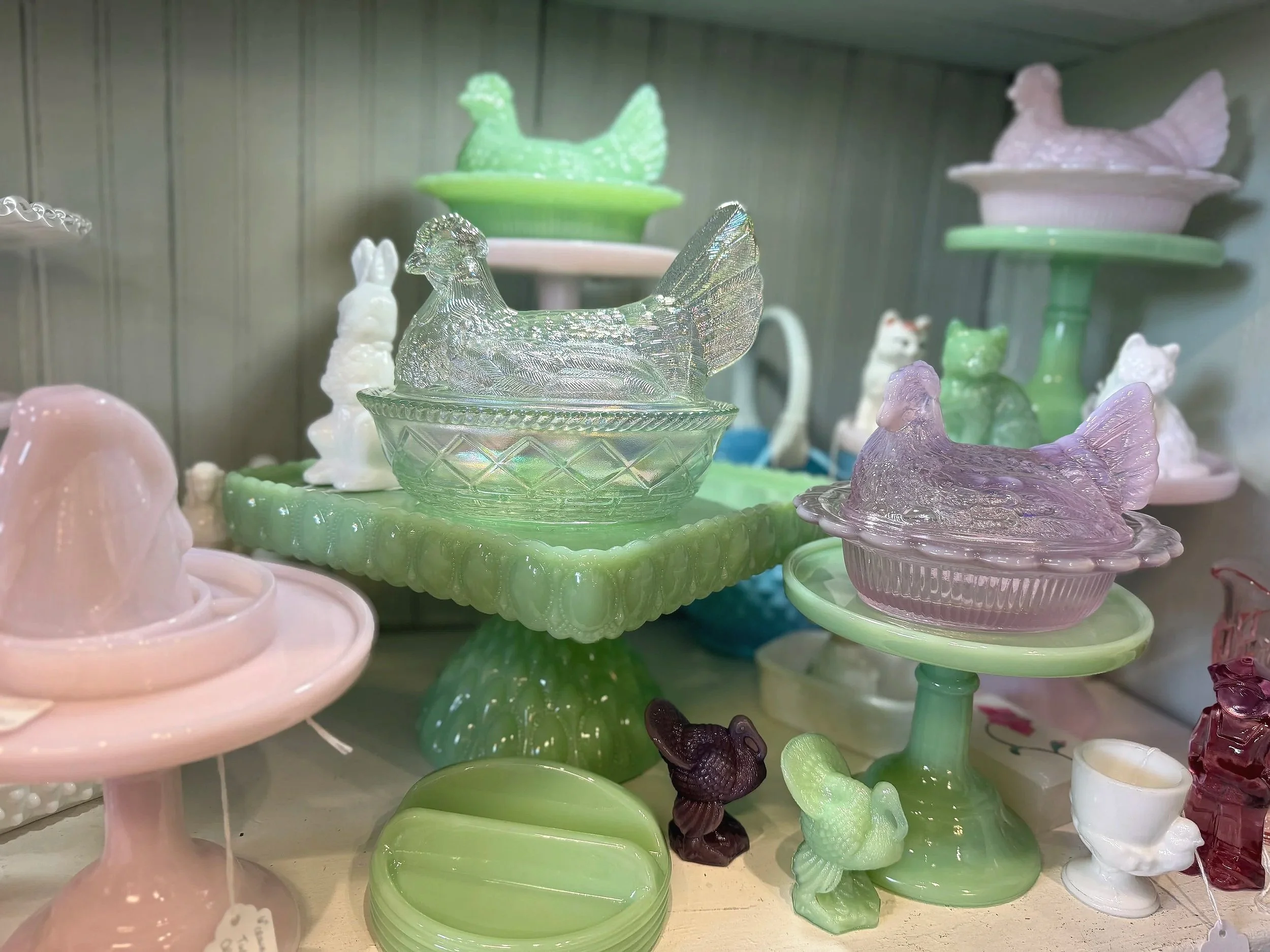Display of colorful glass figurines including chickens, roosters, birds, and other animals on glass stands and trays, with pastel colors like pink, green, purple, and white.