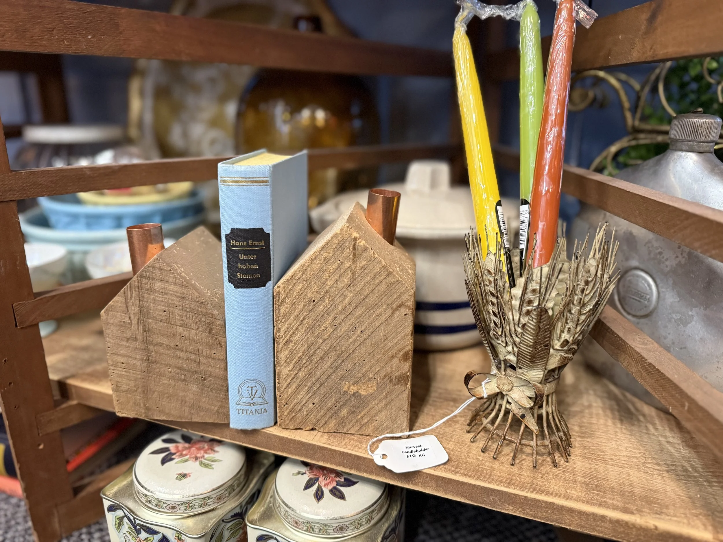 Decorative shelf holding a wooden bookend, a blue book titled 'Hans Ernst Unter hohen Sternen', a ceramic vase, a colorful feathered centerpiece, and some tin containers with floral patterns.