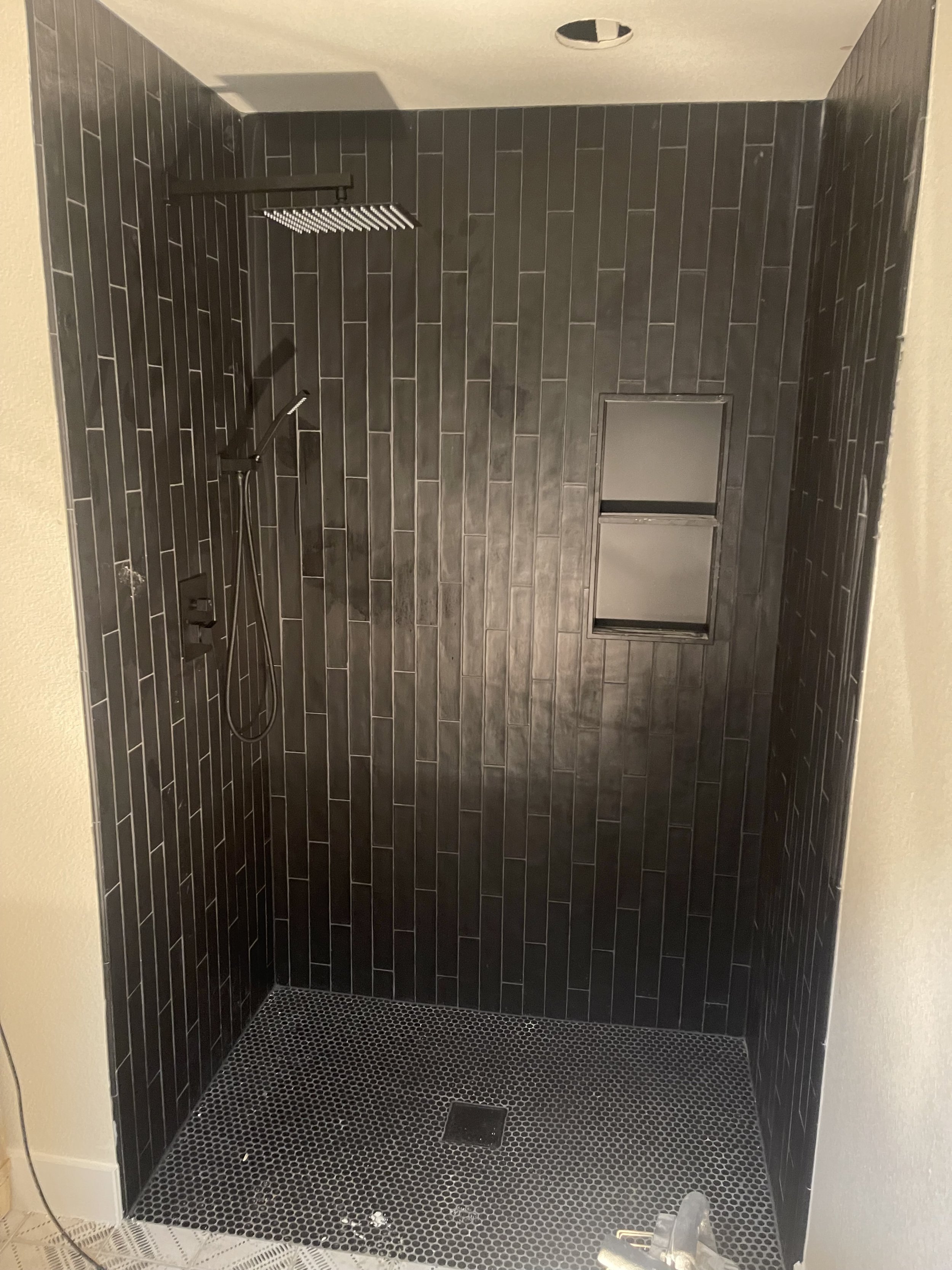 A modern walk-in shower with black tile walls and a black hexagonal tile floor, a ceiling showerhead, a handheld showerhead, built-in shelf, and a drain in the center of the floor.