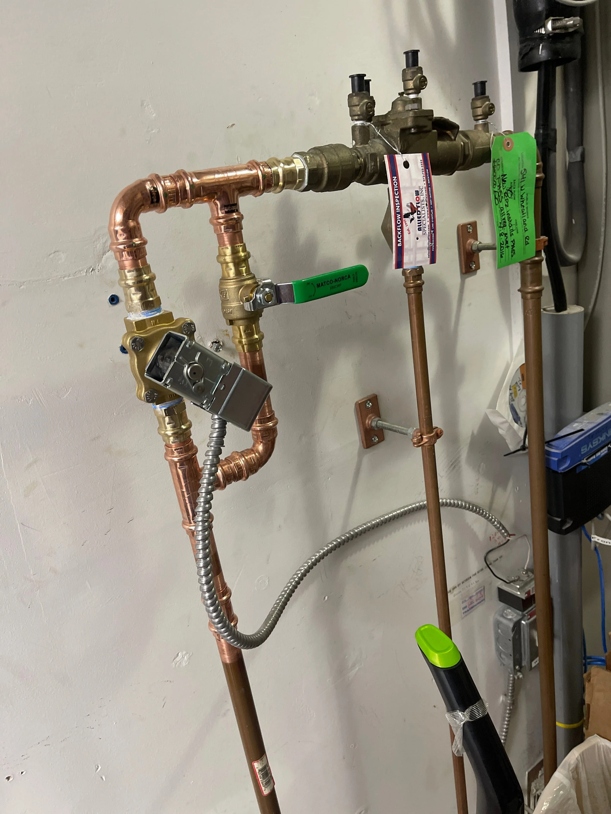 Plumbing system with copper and brass pipes, a valve with a green handle, a pressure gauge, and various tags attached to the pipes, mounted on a white wall.