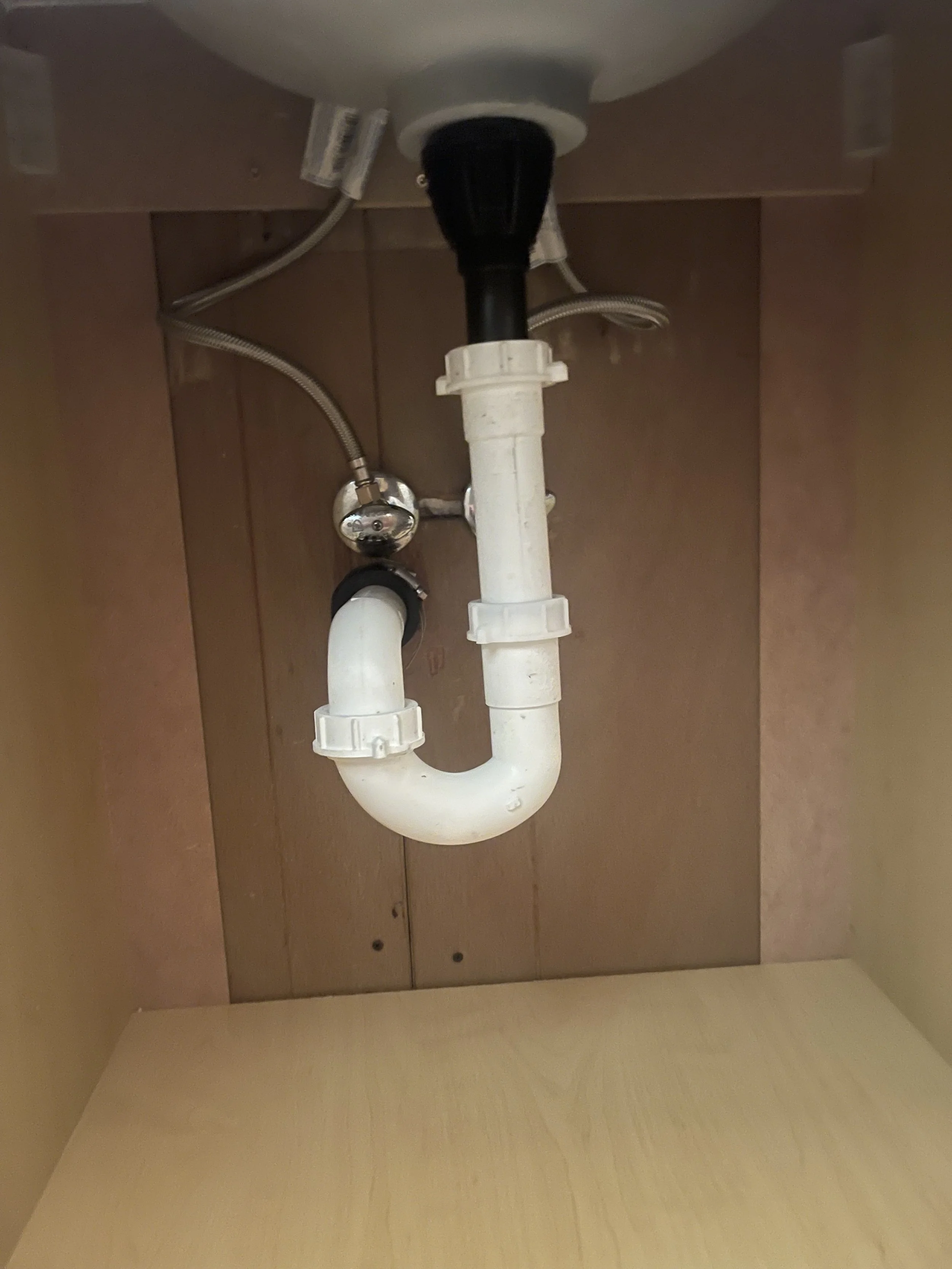 Under the kitchen sink showing a drain pipe, water supply lines, and connections.