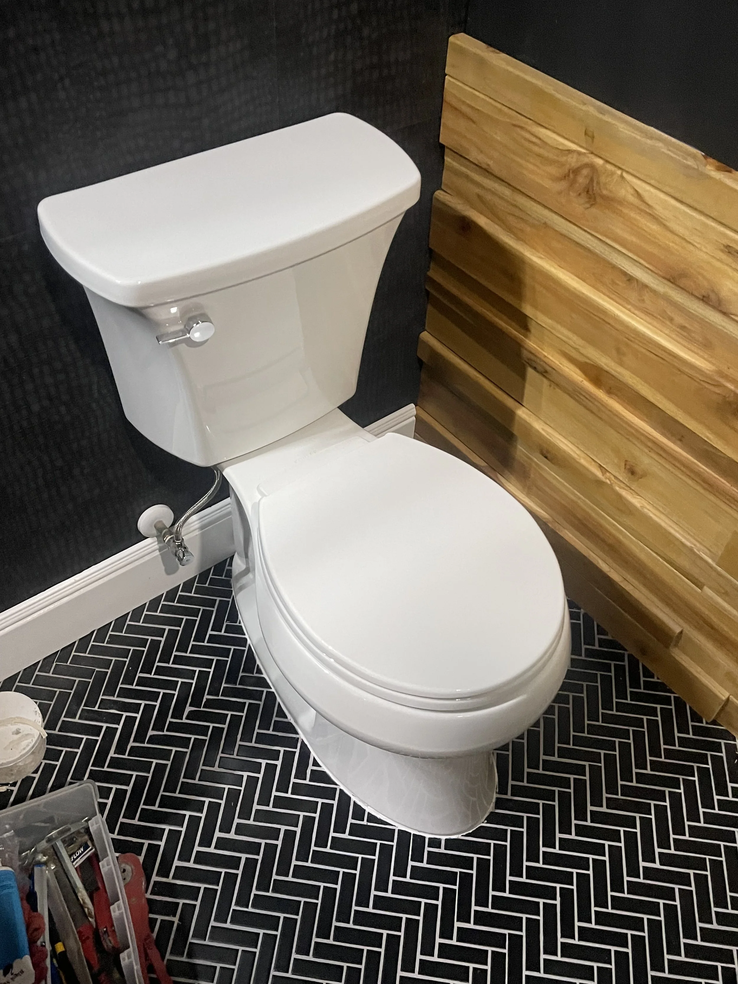 A white toilet with a bidet spray handle mounted on the side, situated in a bathroom with black and white herringbone-patterned floor tiles, a black wall, and a wooden panel on one side.