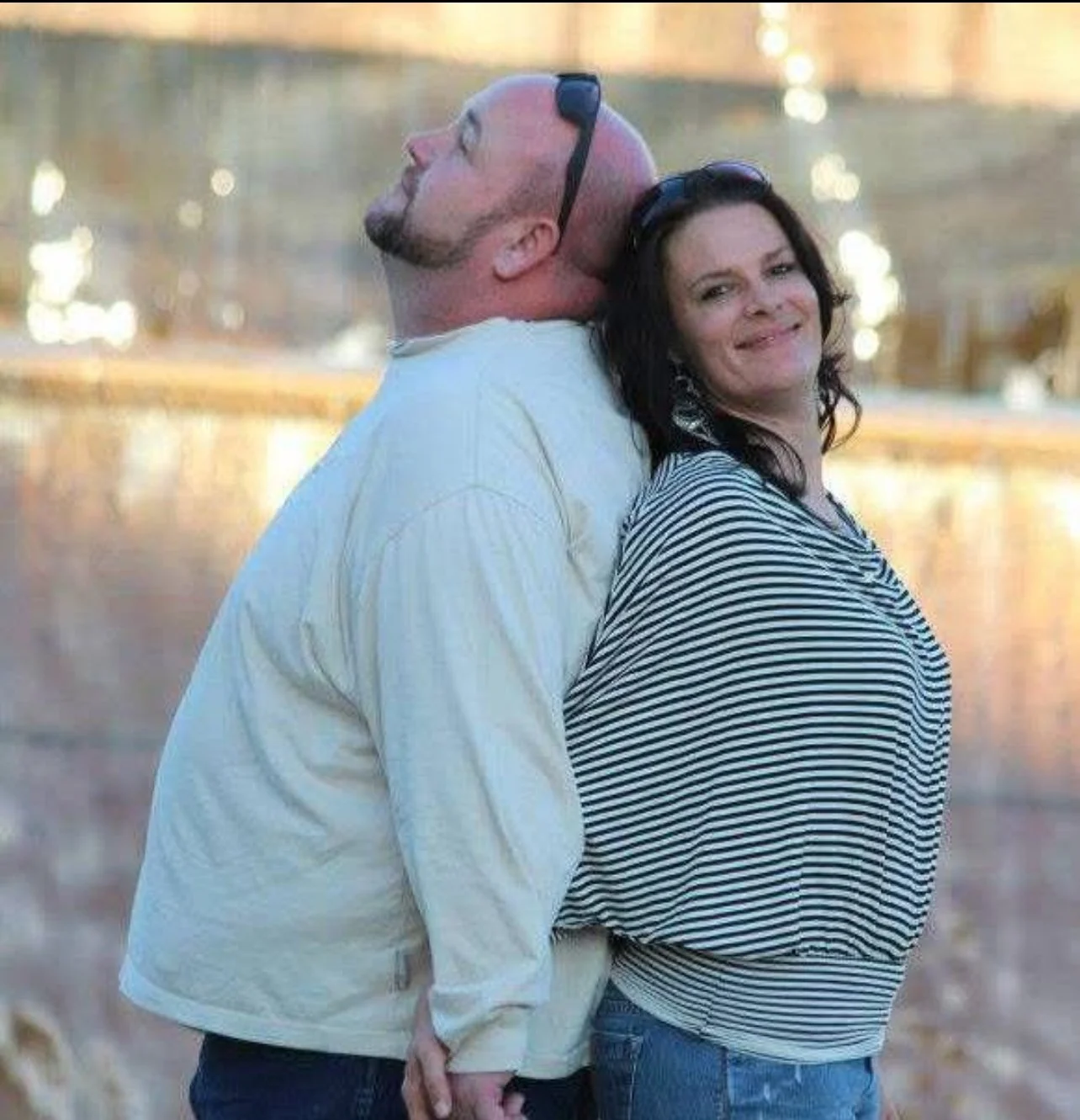 A man and woman standing outdoors near a body of water, back to back, with the man leaning his head on the woman's shoulder. The man is bald with sunglasses on his head, wearing a cream-colored shirt. The woman has dark, curly hair, wearing a striped shirt and smiling.