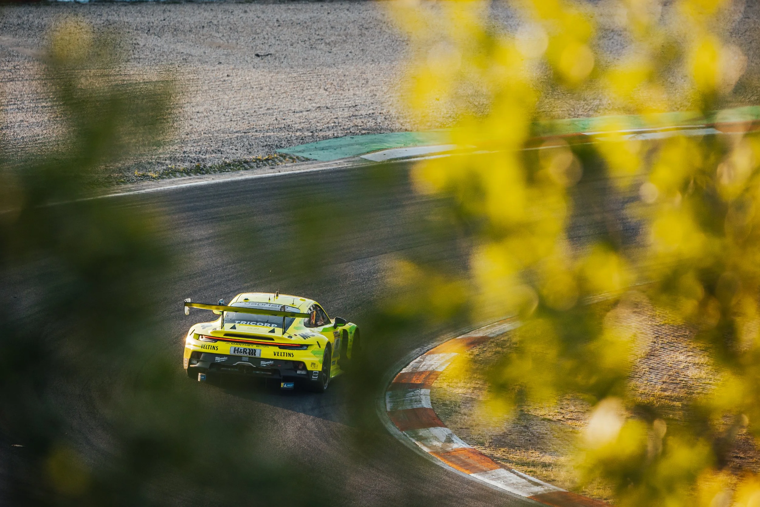 A yellow race car with sponsor logos driving on a curving race track, partially obscured by blurred yellow and green foliage in the foreground.