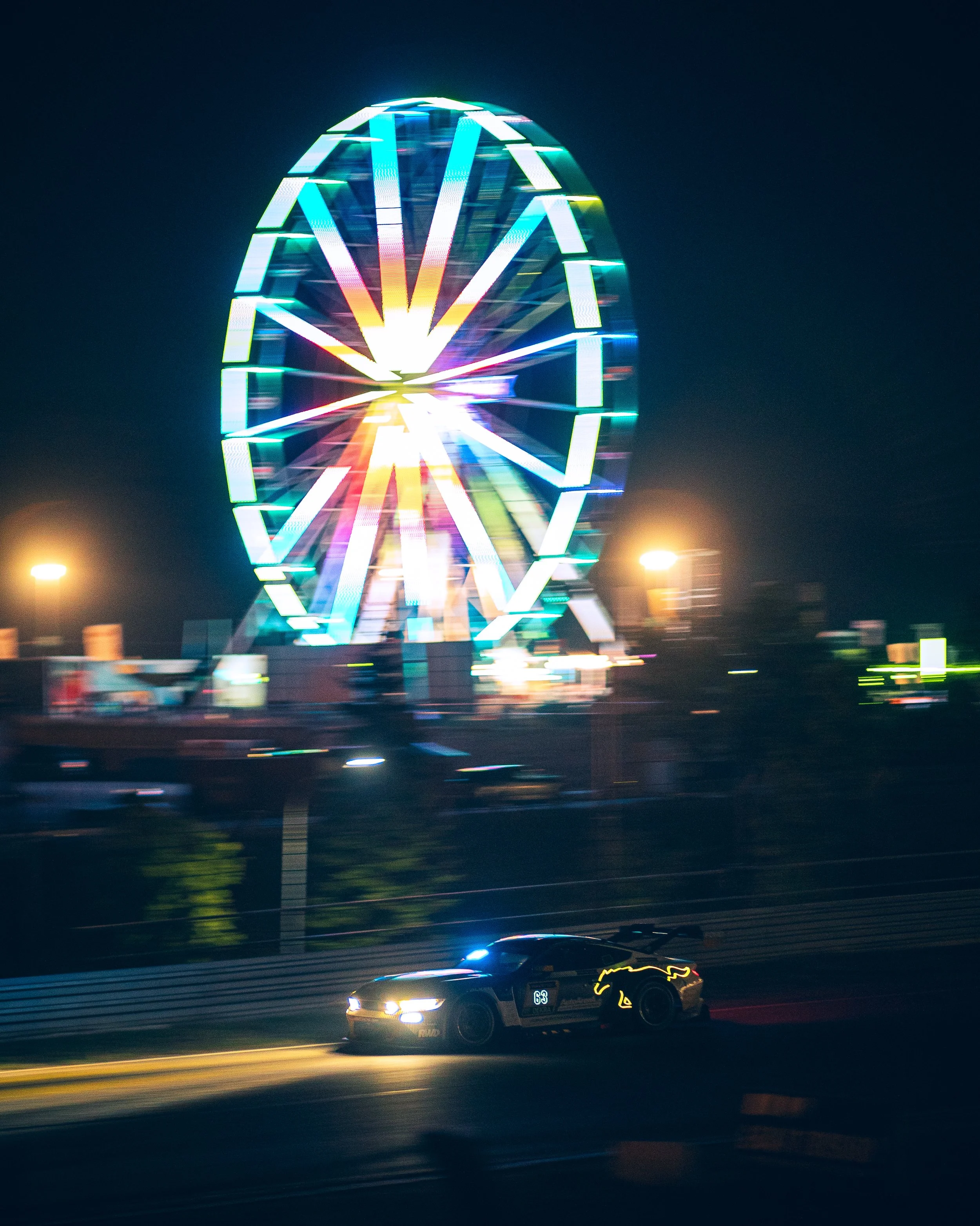 A race car moving on a track at night with a large, brightly lit Ferris wheel in the background.