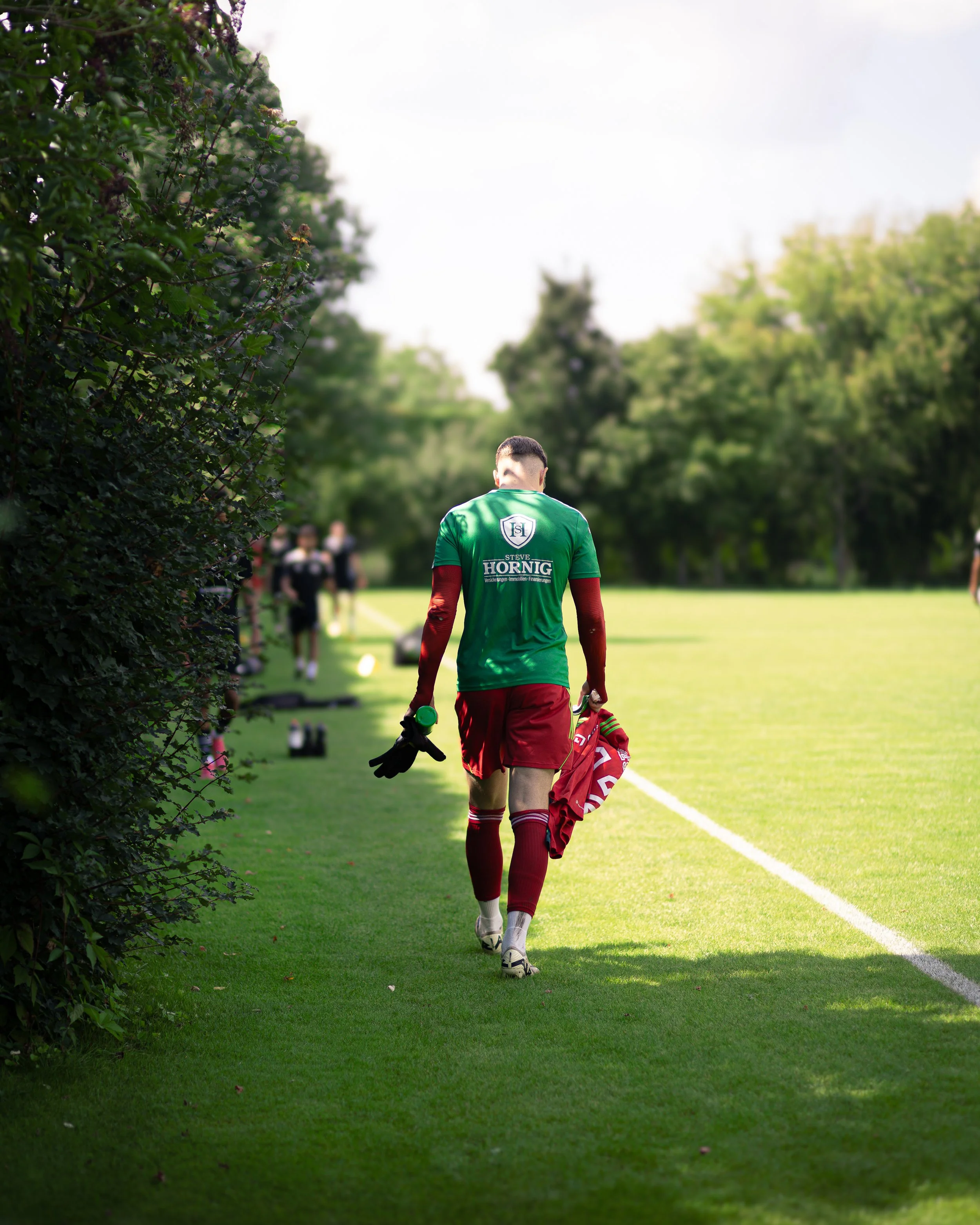 A soccer player walking away on a grassy field holding a red jacket and gloves, with teammates in the background.