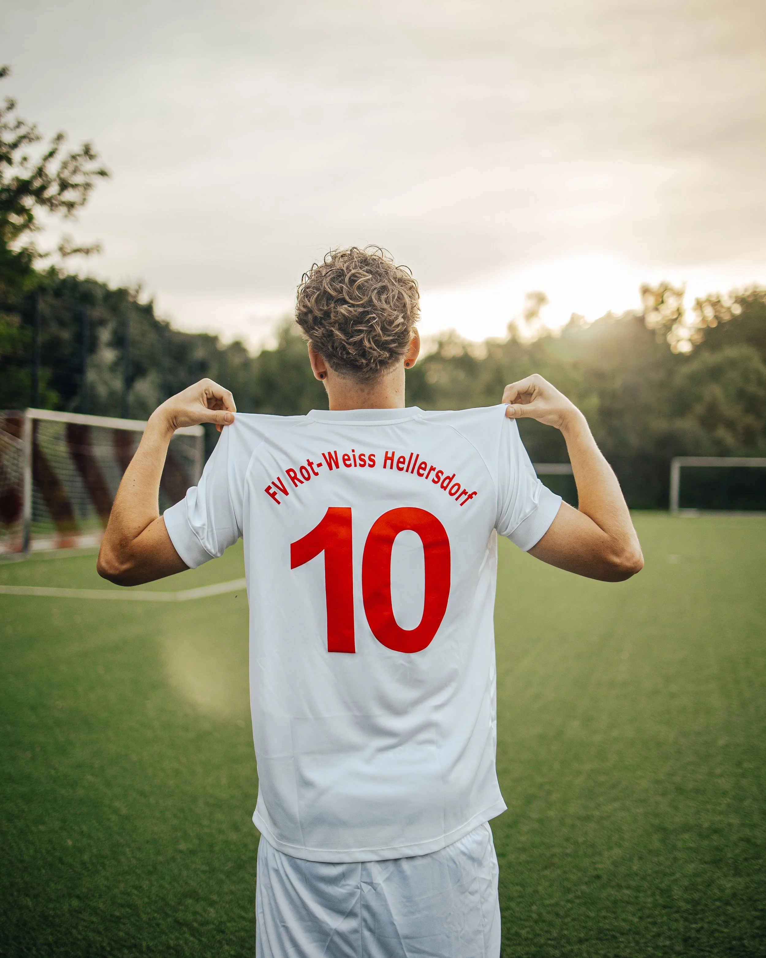 A person with curly hair holding up a white soccer jersey with red text and number '10' on the back, standing on a soccer field at sunset.