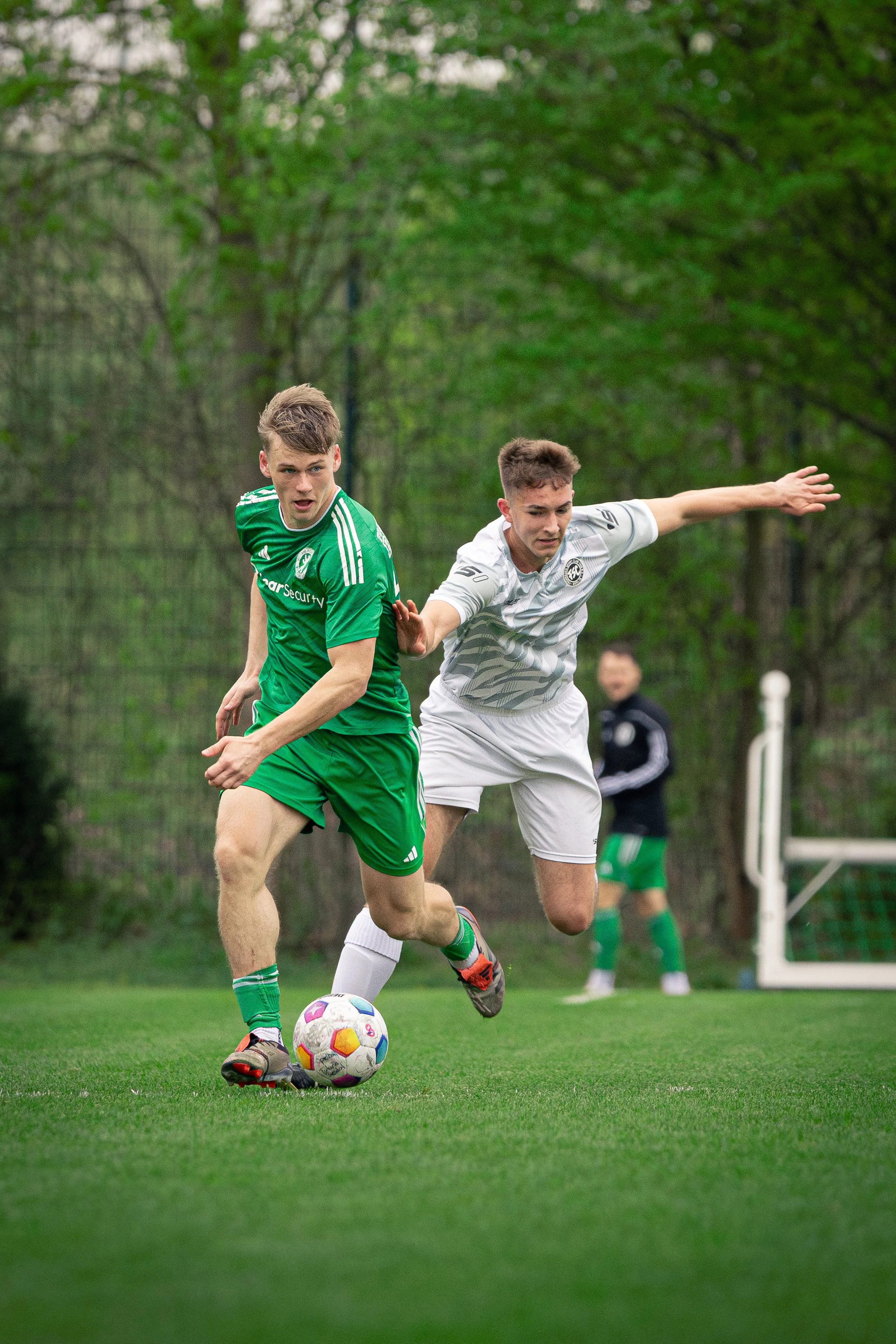 Two young male soccer players competing for the ball during a match on a grassy field with trees in the background. One is wearing a green uniform, and the other is wearing a white uniform.