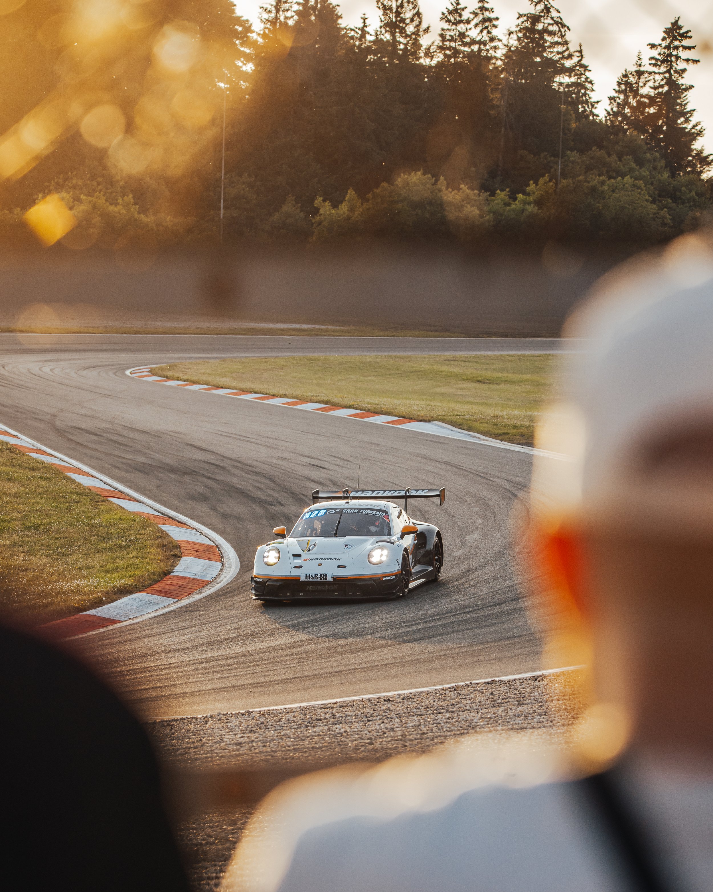 A race car driving on a curved race track during sunset, viewed through a blurry foreground with trees in the background.