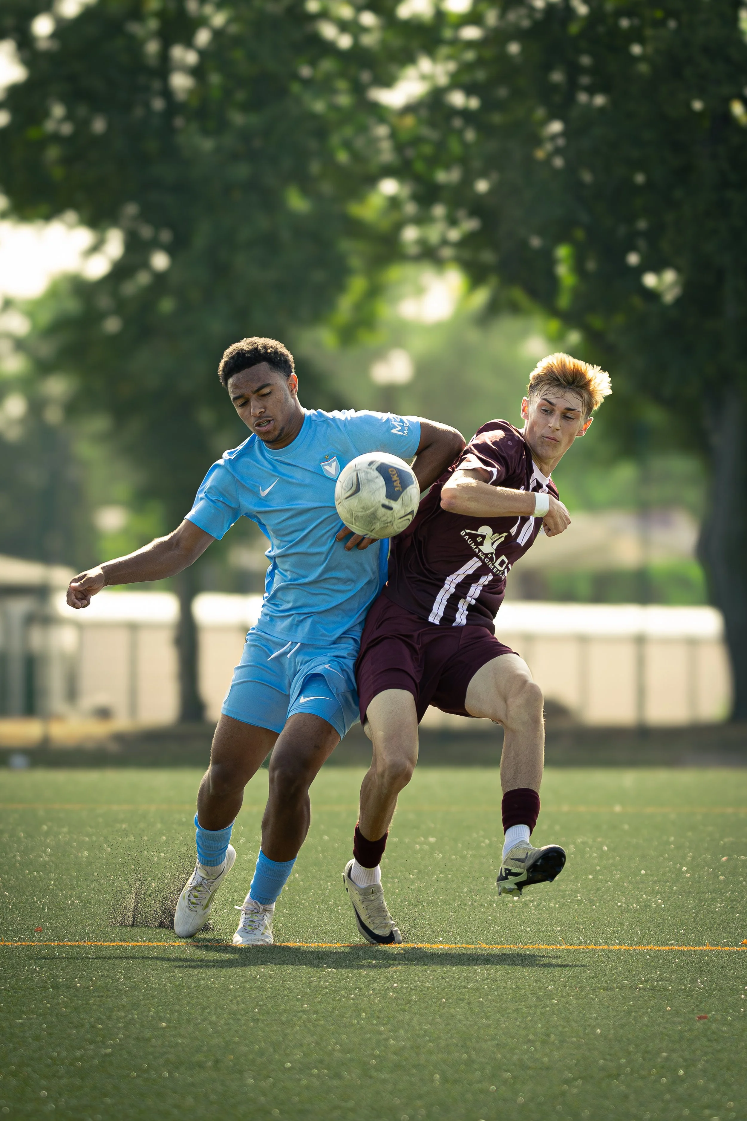 Two soccer players, one in a blue uniform and the other in a maroon uniform, competing for the ball on a grassy field with trees in the background.
