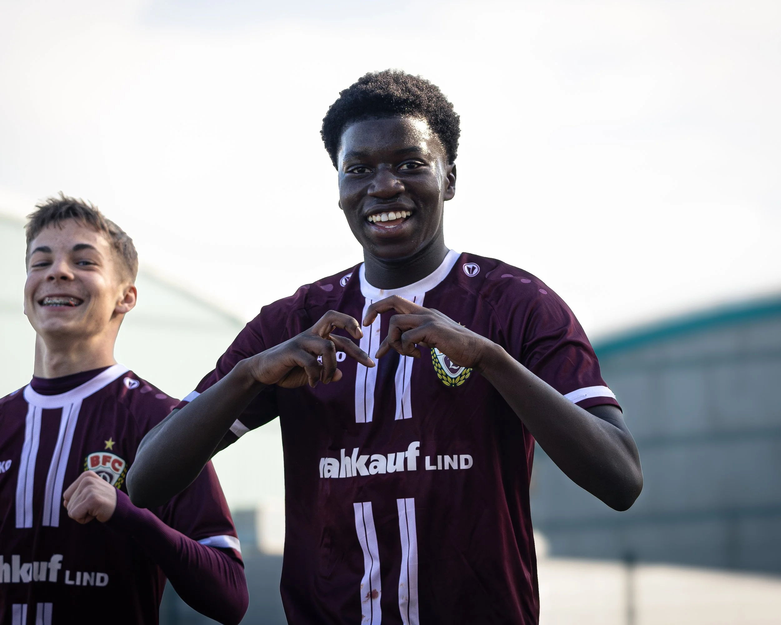 Two soccer players in maroon jerseys celebrating, with one smiling and making a heart gesture with his fingers, outdoors during daylight.