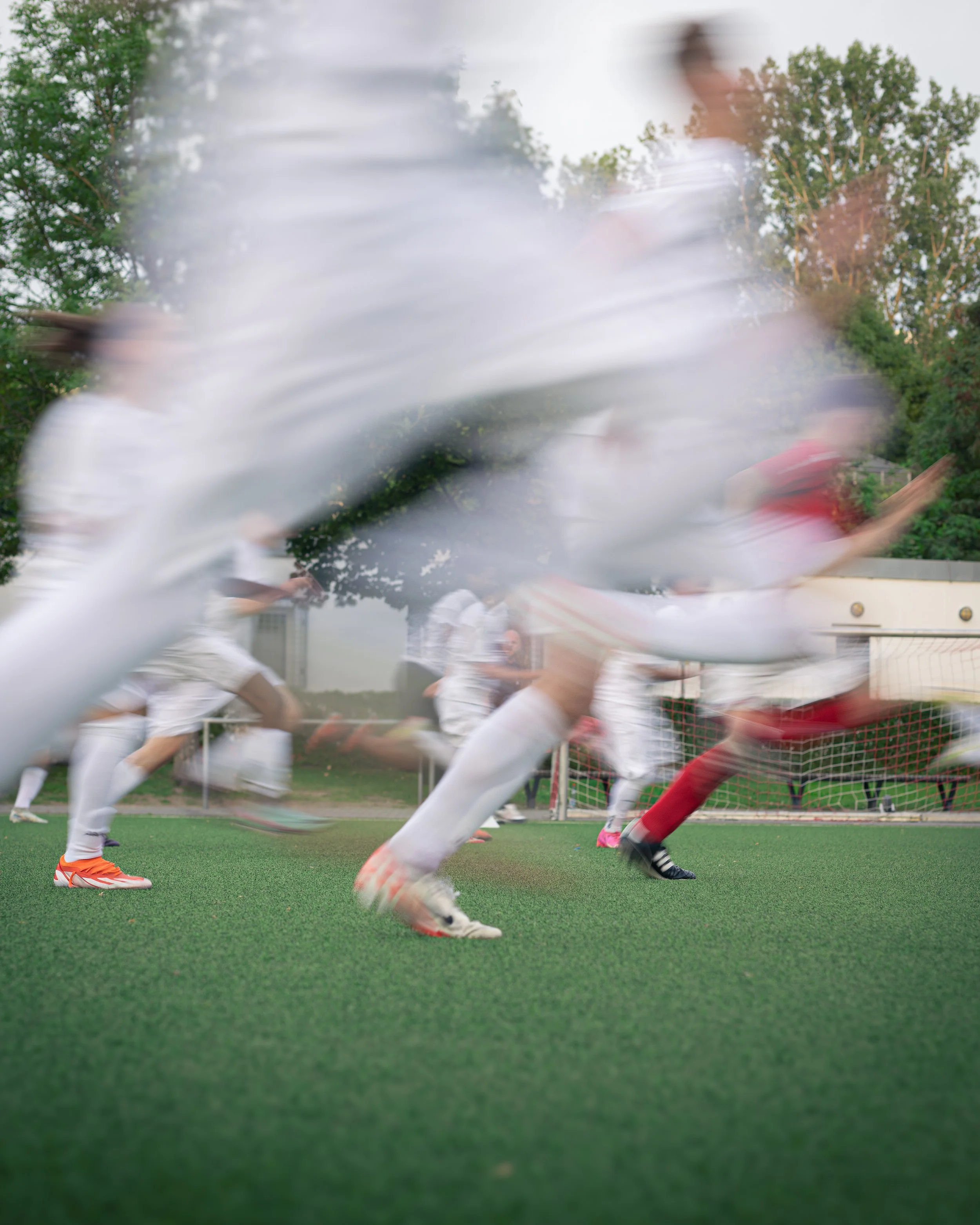 Blurry motion shot of soccer players running on a field, wearing white uniforms with one player in red, with trees and a goalpost in the background.