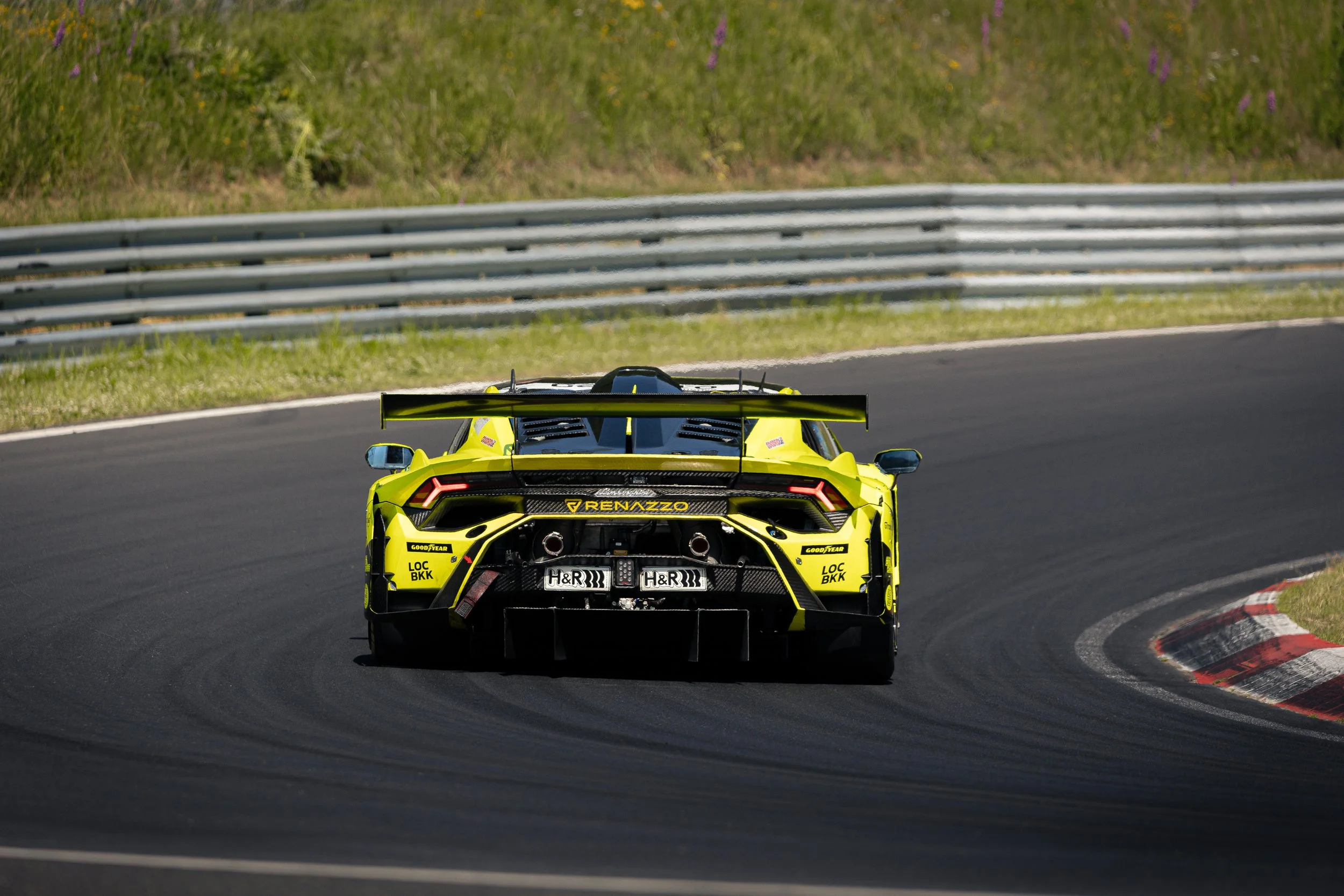 A bright yellow race car with a rear spoiler driving on a racetrack, with safety barriers and grass in the background.