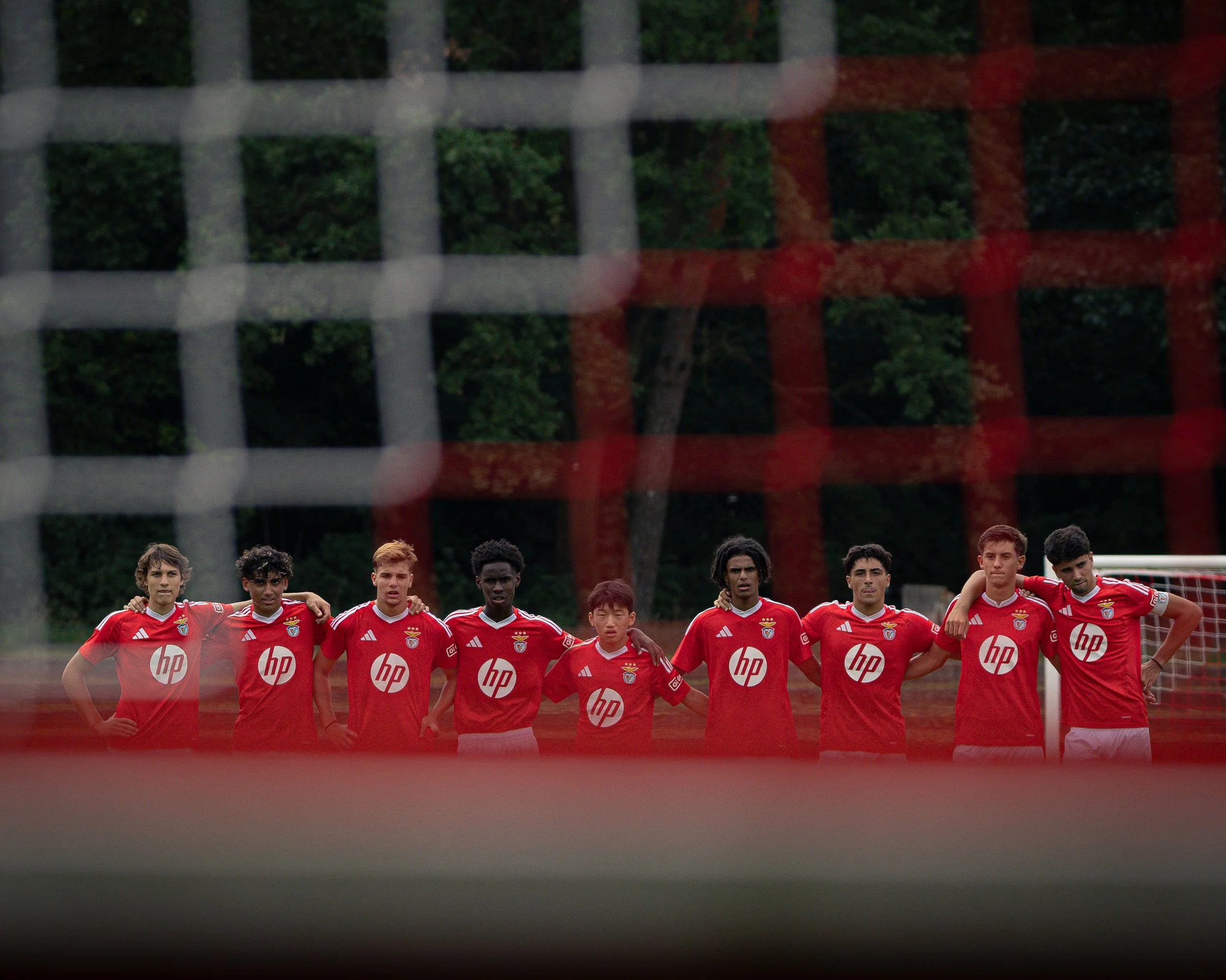 A group of nine soccer players in red uniforms standing in a line on a soccer field, with a goal and trees in the background, viewed through a red and white net.