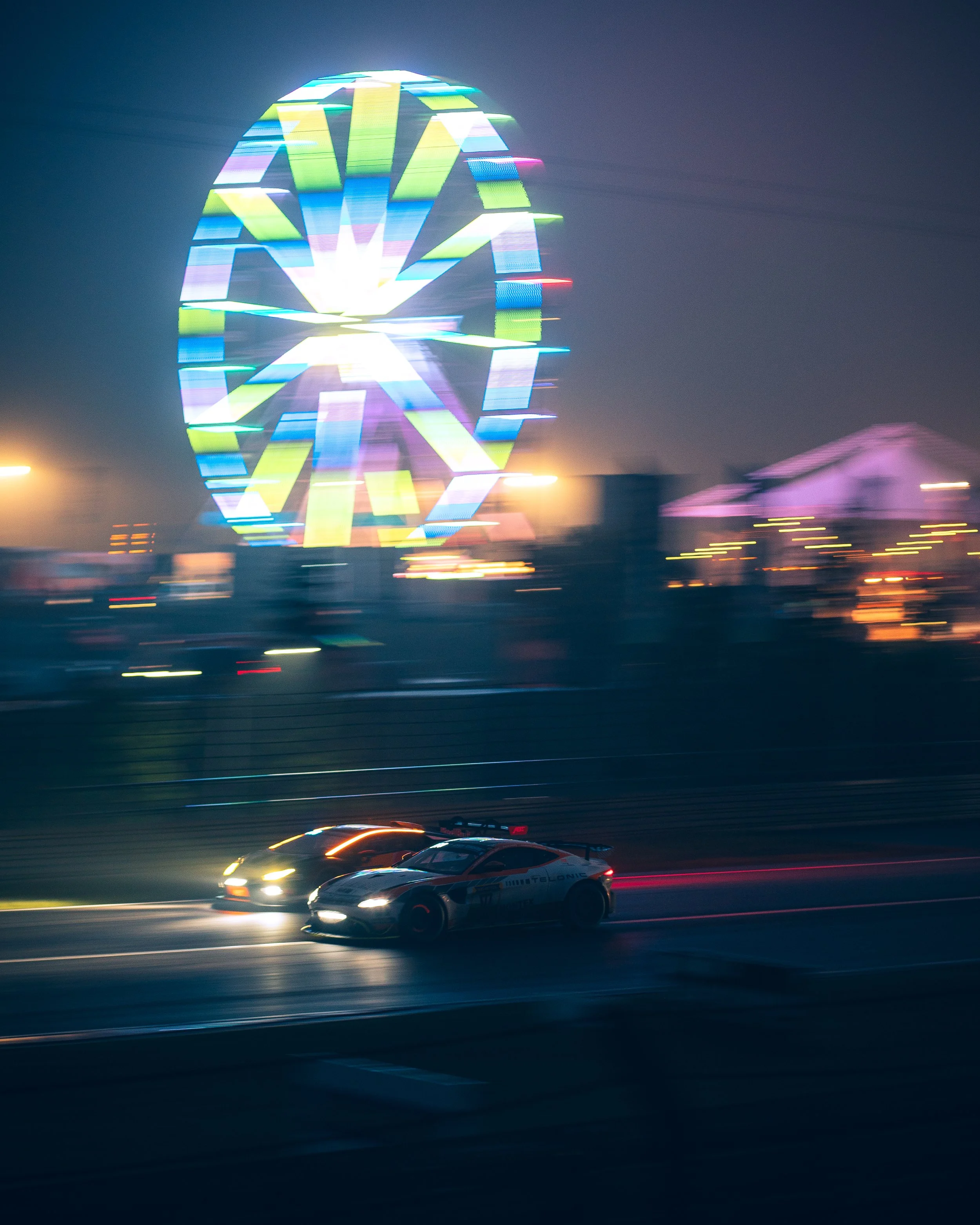 Nighttime scene of a race car on a track with a brightly lit Ferris wheel in the background, blurry movement indicating speed.