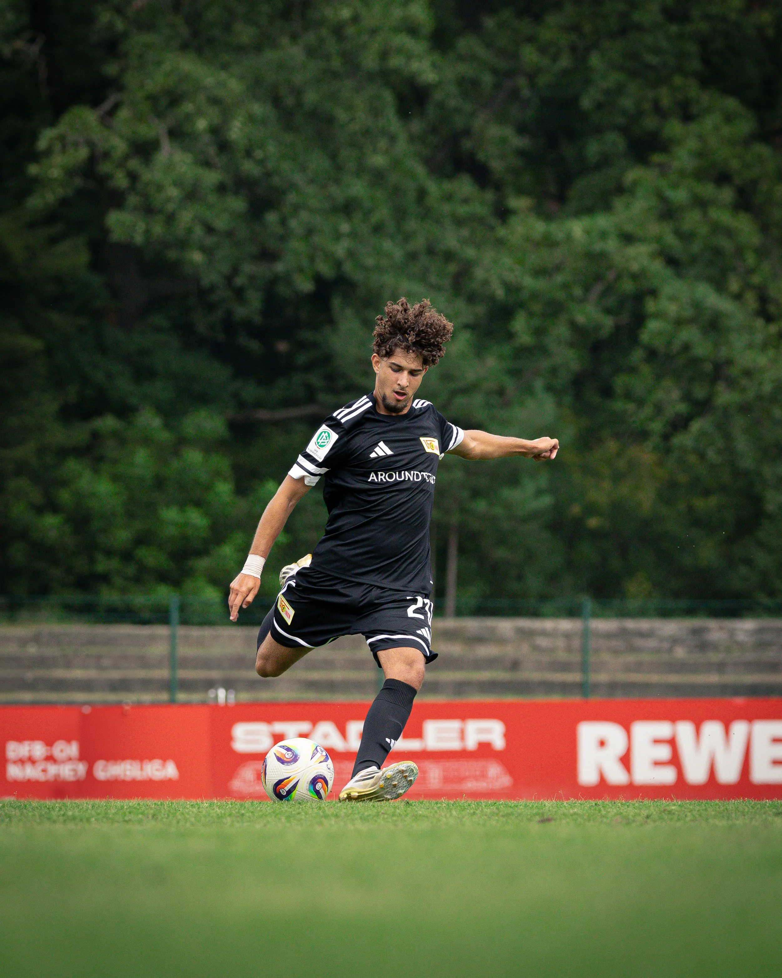 A soccer player in a black uniform kicking a soccer ball on a field with green trees in the background.