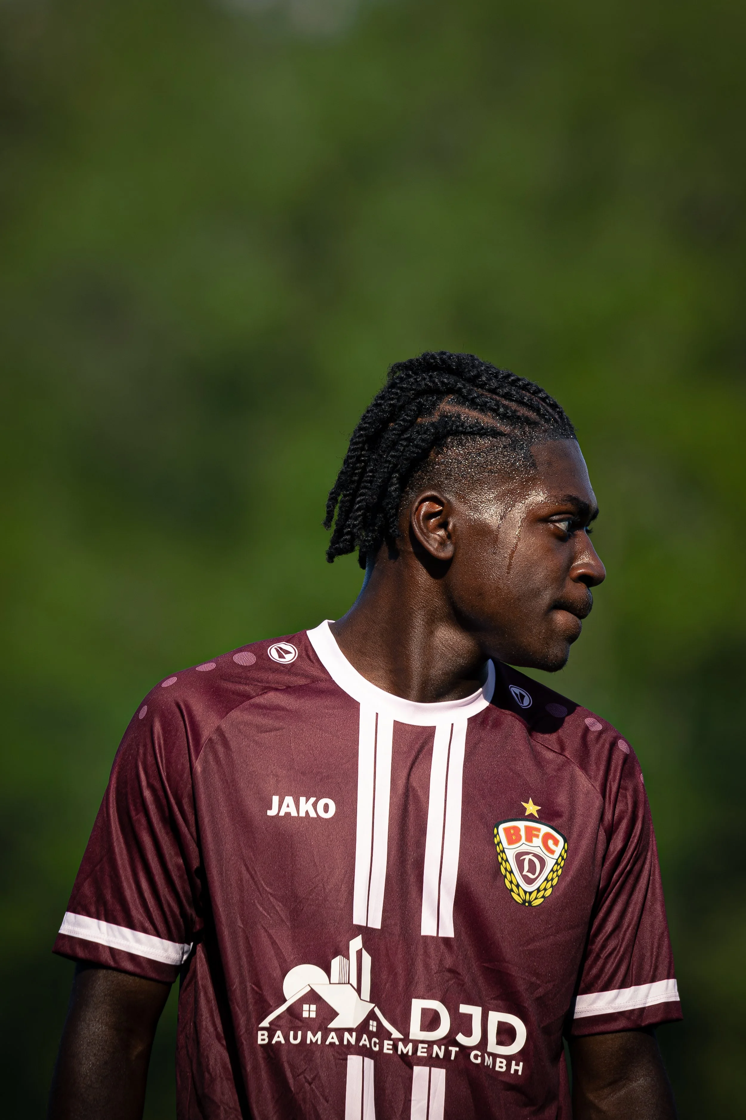 A male soccer player with dreadlocks, wearing a maroon jersey with white accents, standing outdoors with a blurred green background.
