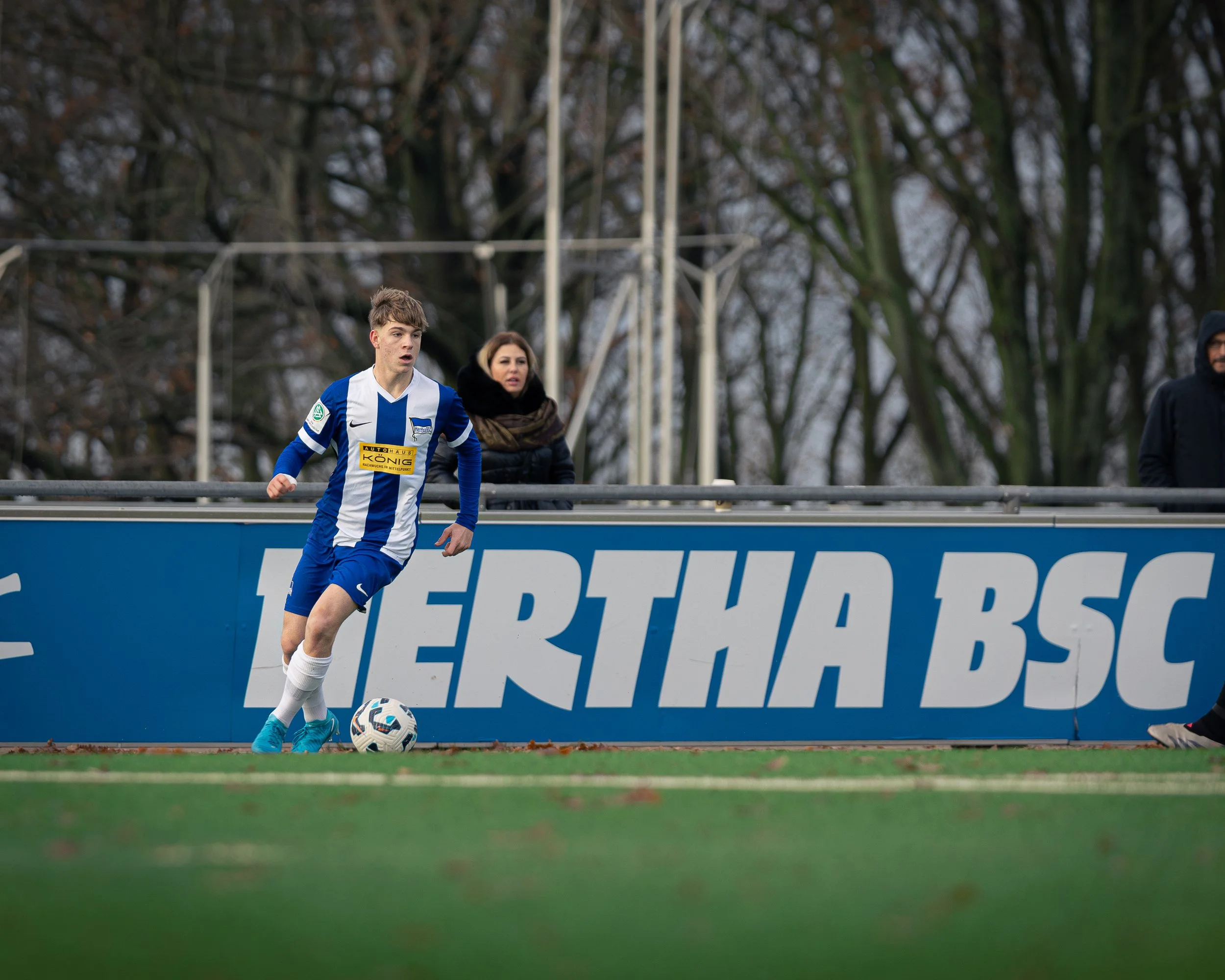 A soccer player in a blue and white uniform on the field, with a woman and a man watching behind a barrier, and trees in the background.