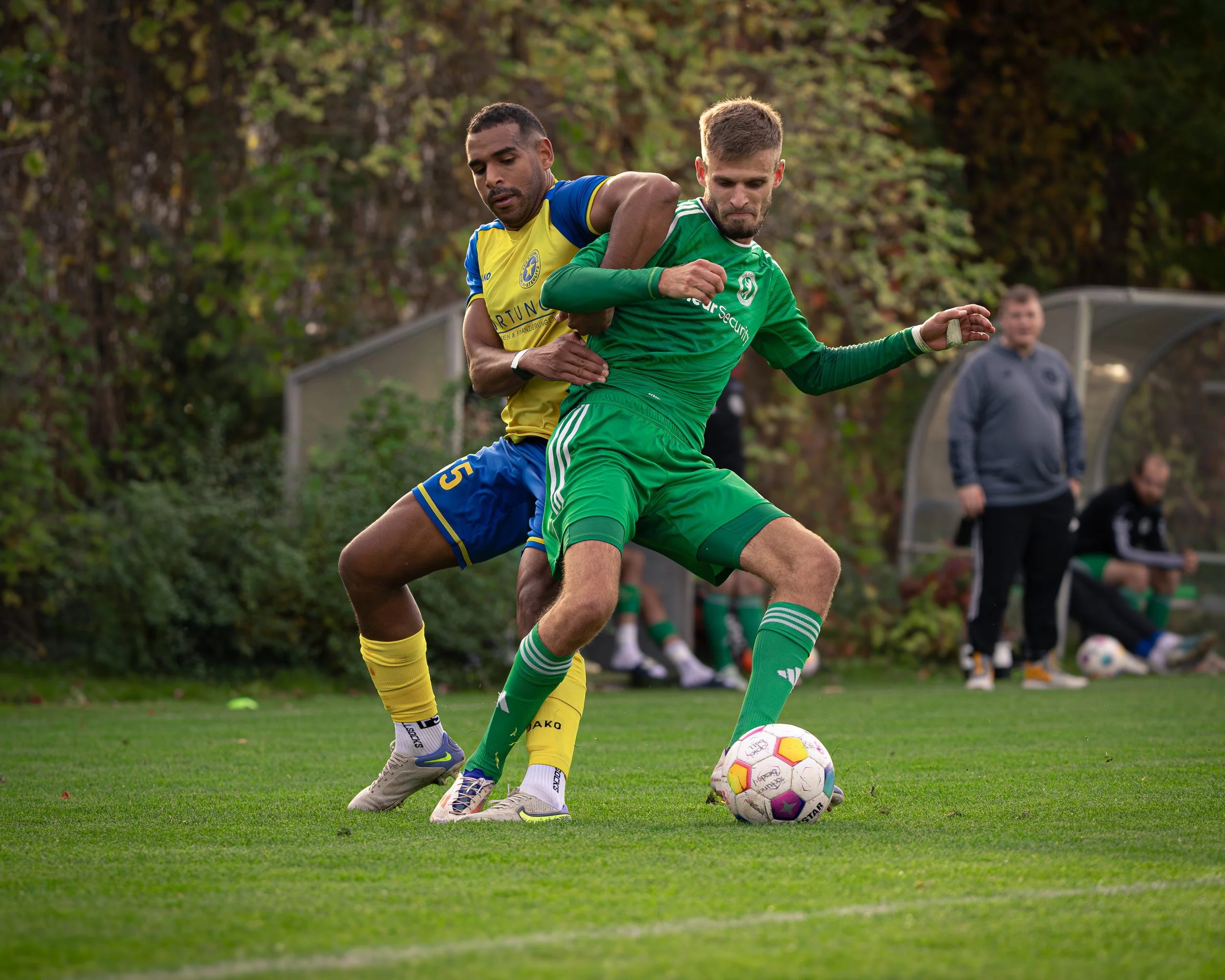 Two soccer players are competing for the ball on a grass field, with players on the bench and a coach or referee in the background, in an outdoor setting with trees.
