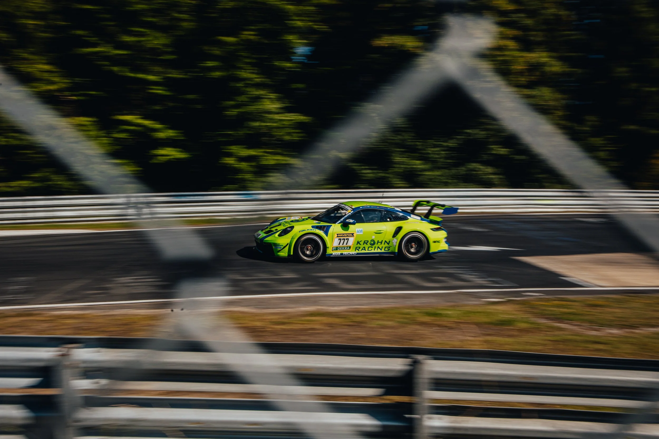 A bright yellow and green race car driving on a race track, viewed through a blurred chain-link fence, with trees in the background.