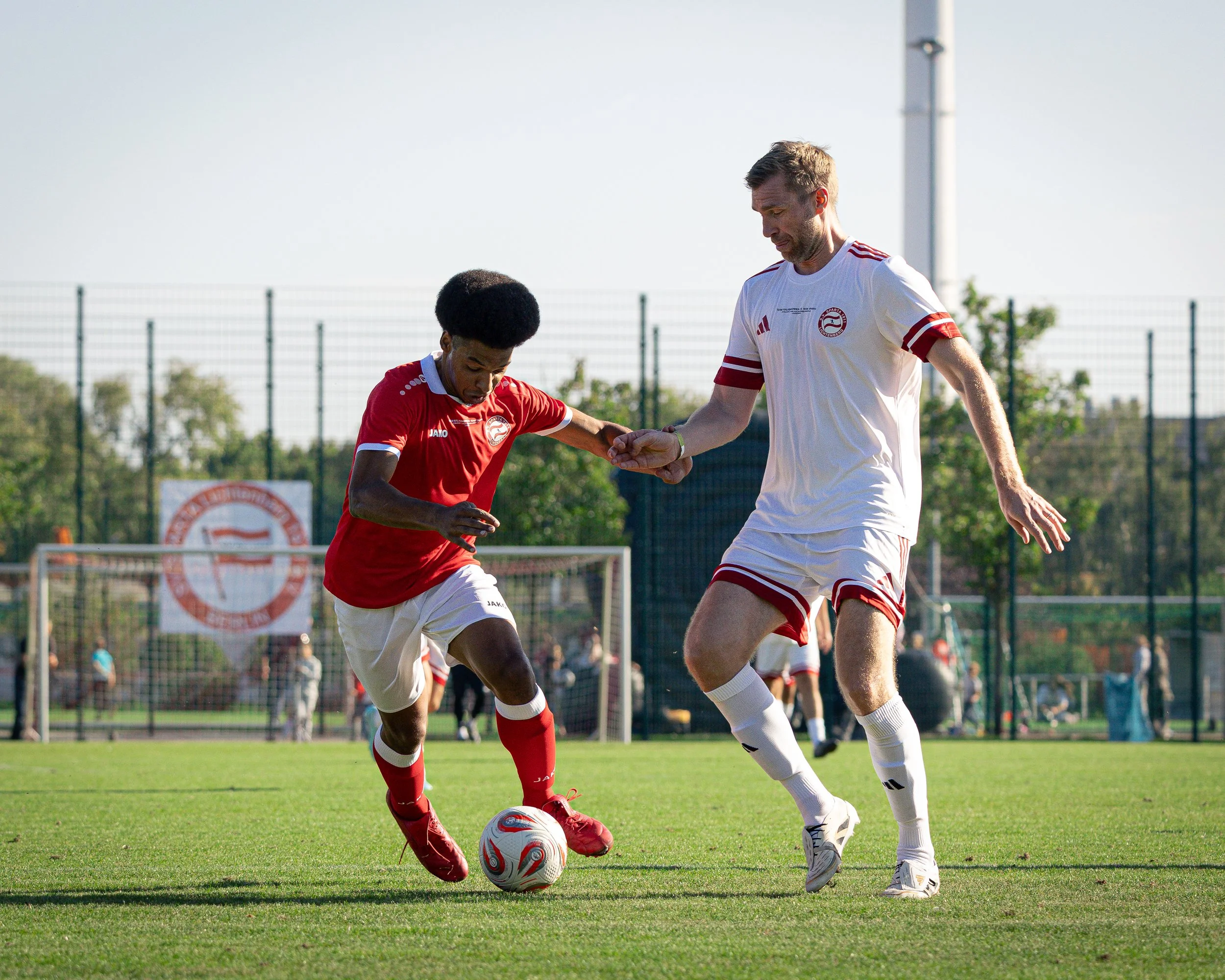 Two soccer players, one in a red jersey and the other in a white jersey, compete for the ball on a soccer field during a game.