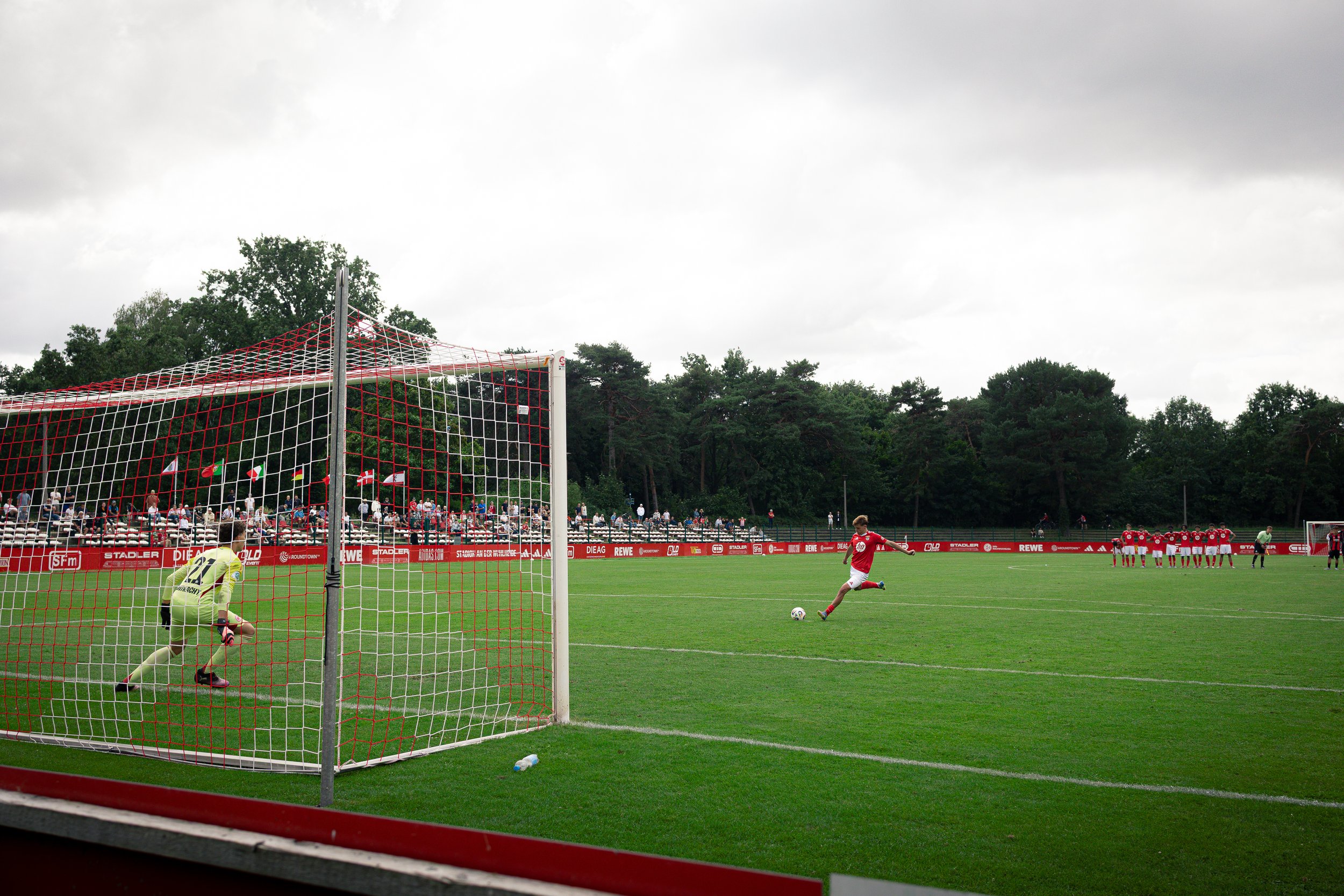 A soccer player takes a penalty kick on a field while the goalkeeper prepares to save. A group of players stands in the background near the goal on a cloudy day.