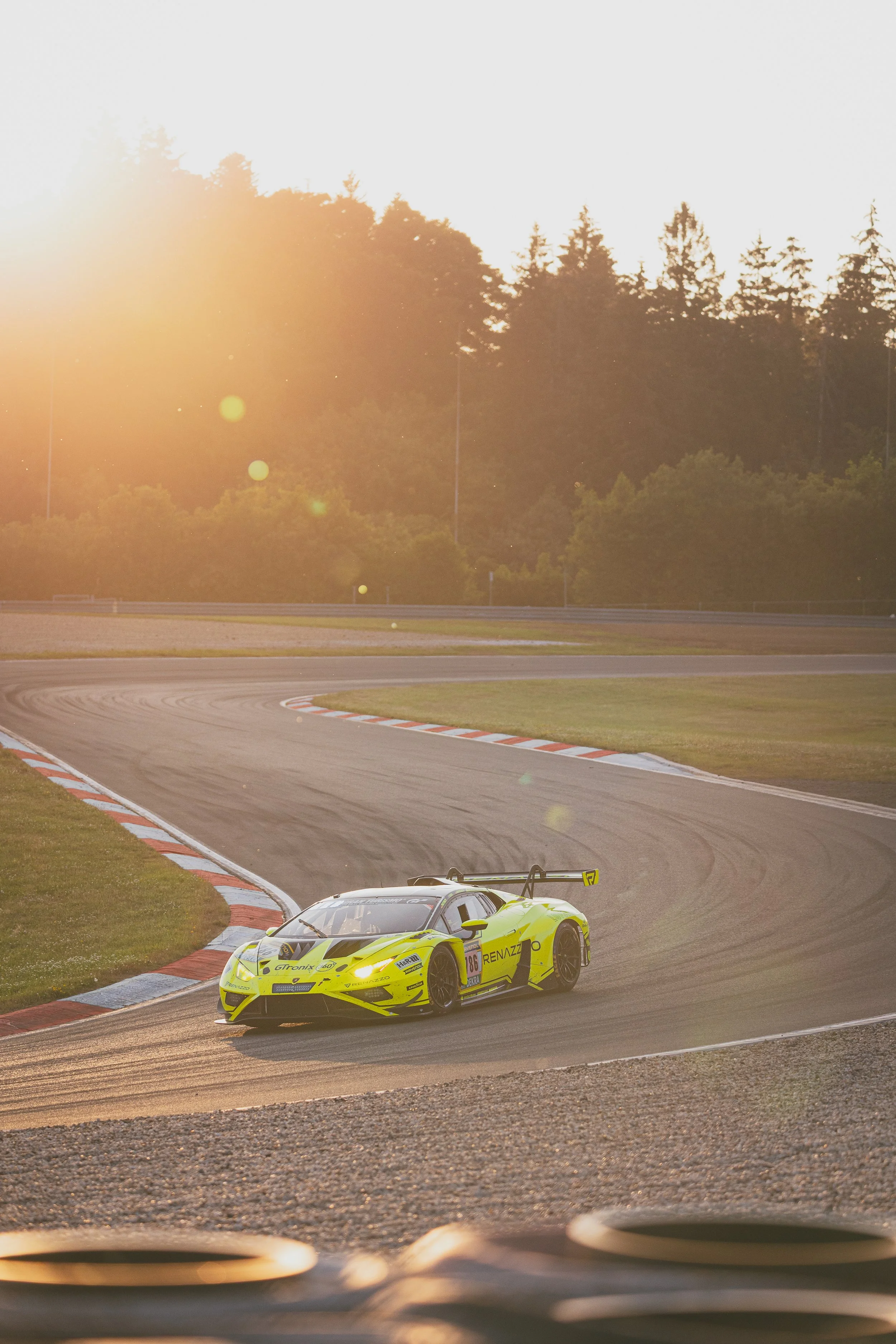 A bright yellow race car speeding on a curving race track at sunset, with the sun shining through trees in the background.