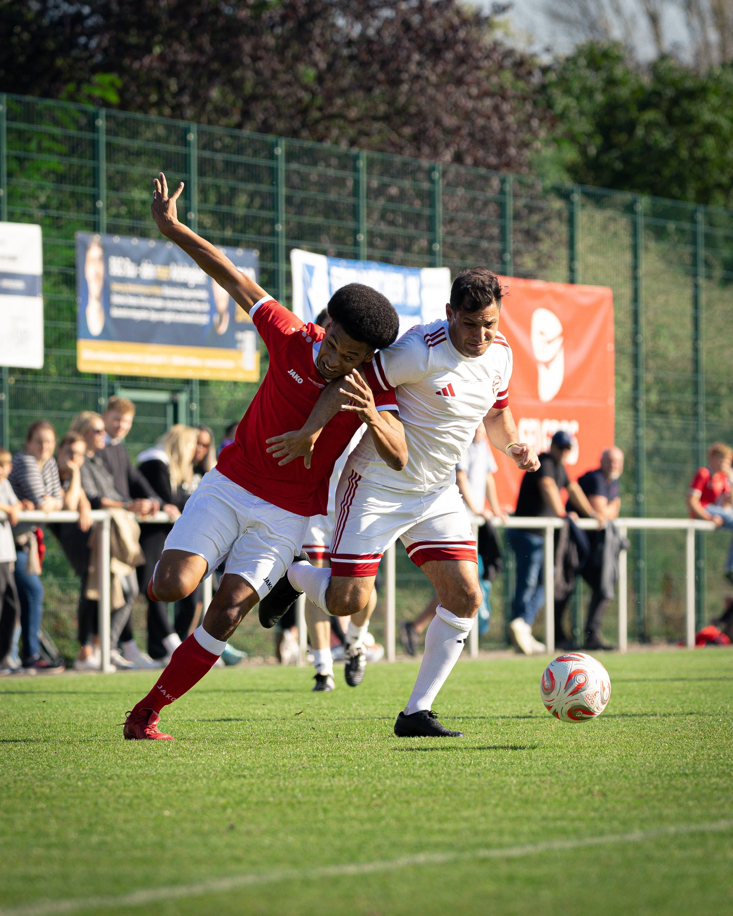 Two soccer players competing for the ball during a match on a grass field, with spectators watching from behind a fence.