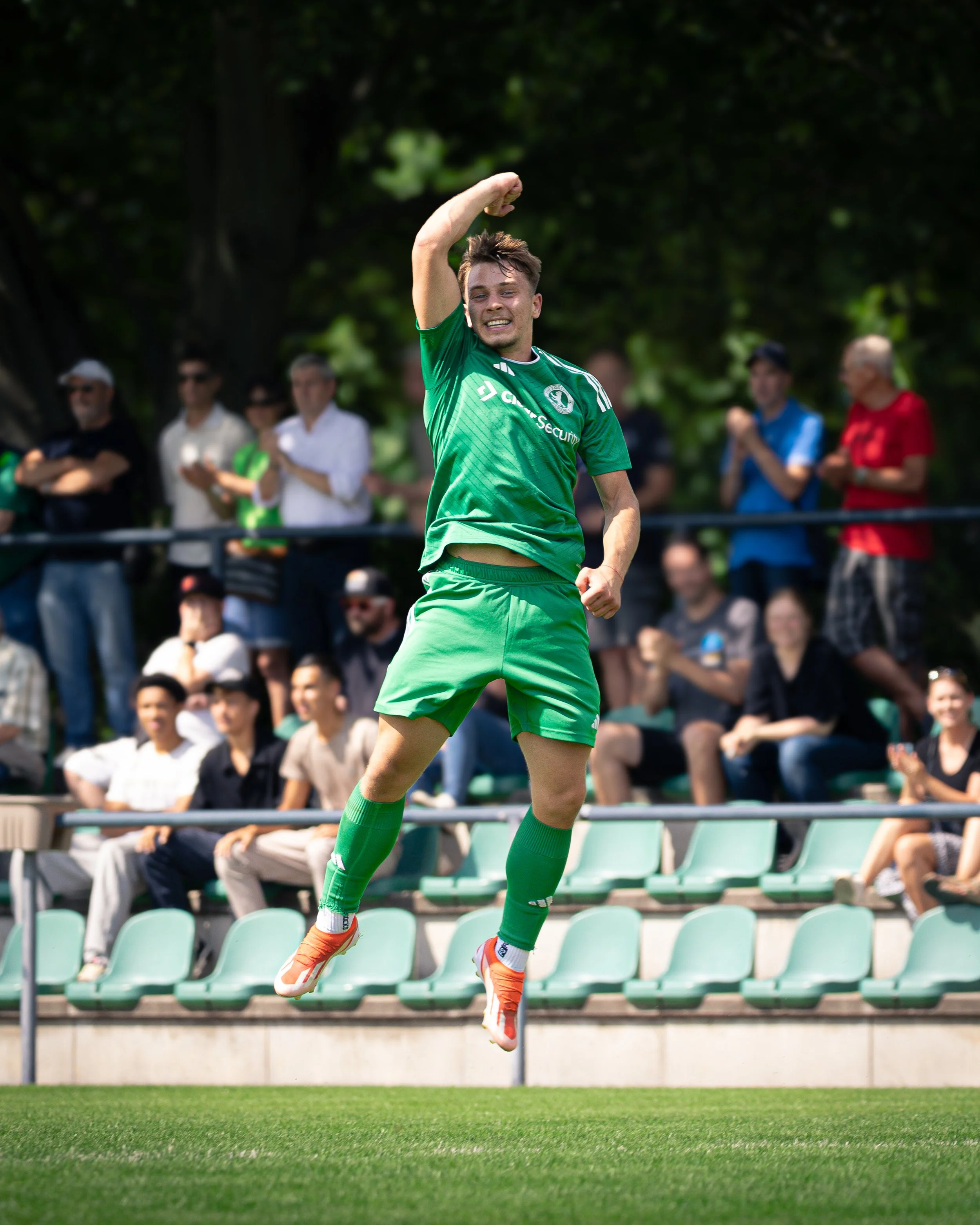 A soccer player in a green uniform jumping in the air on a soccer field during a game, with spectators sitting in the background.