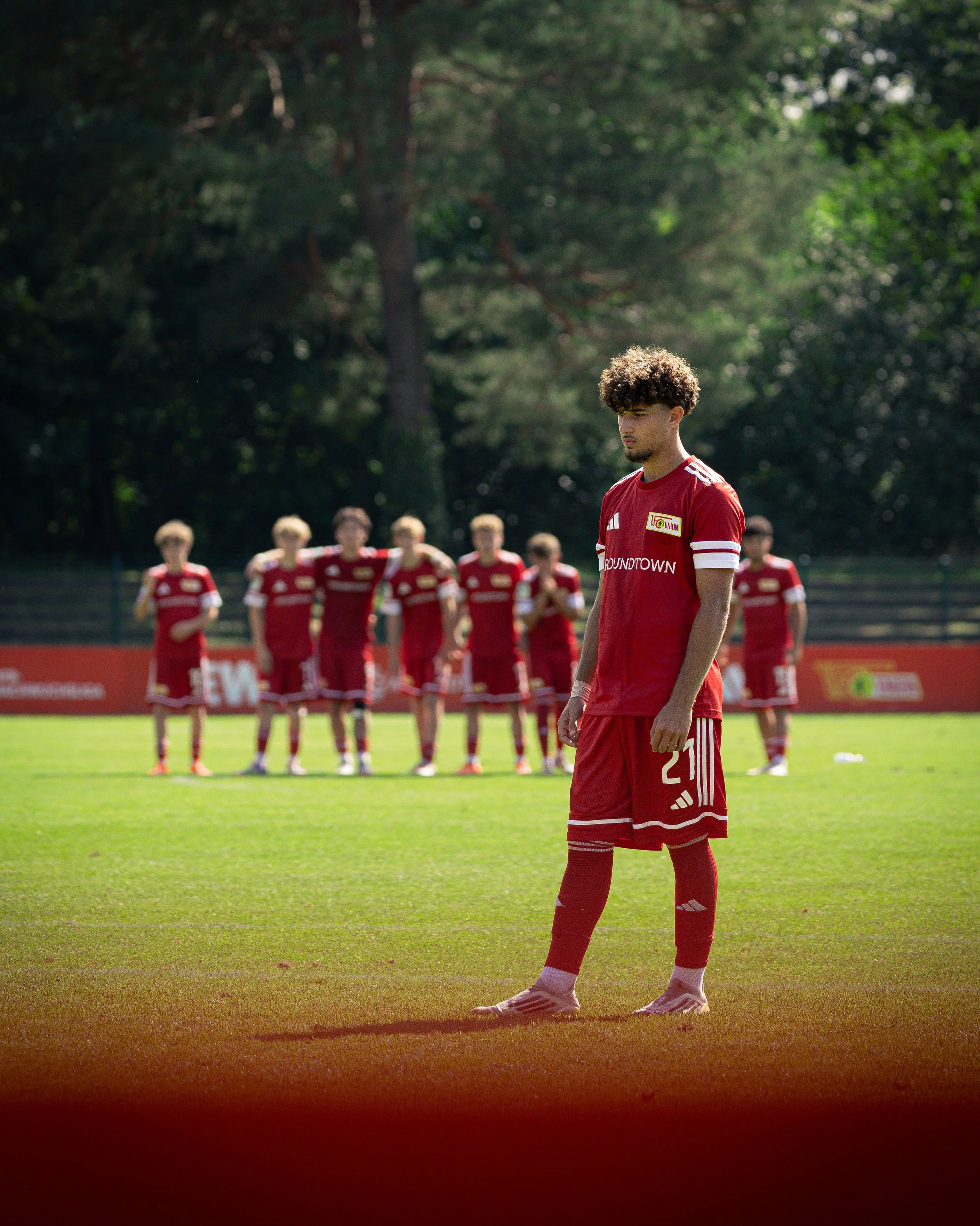 A group of soccer players in red uniforms on a field, with one player in front looking down and others blurred in the background.