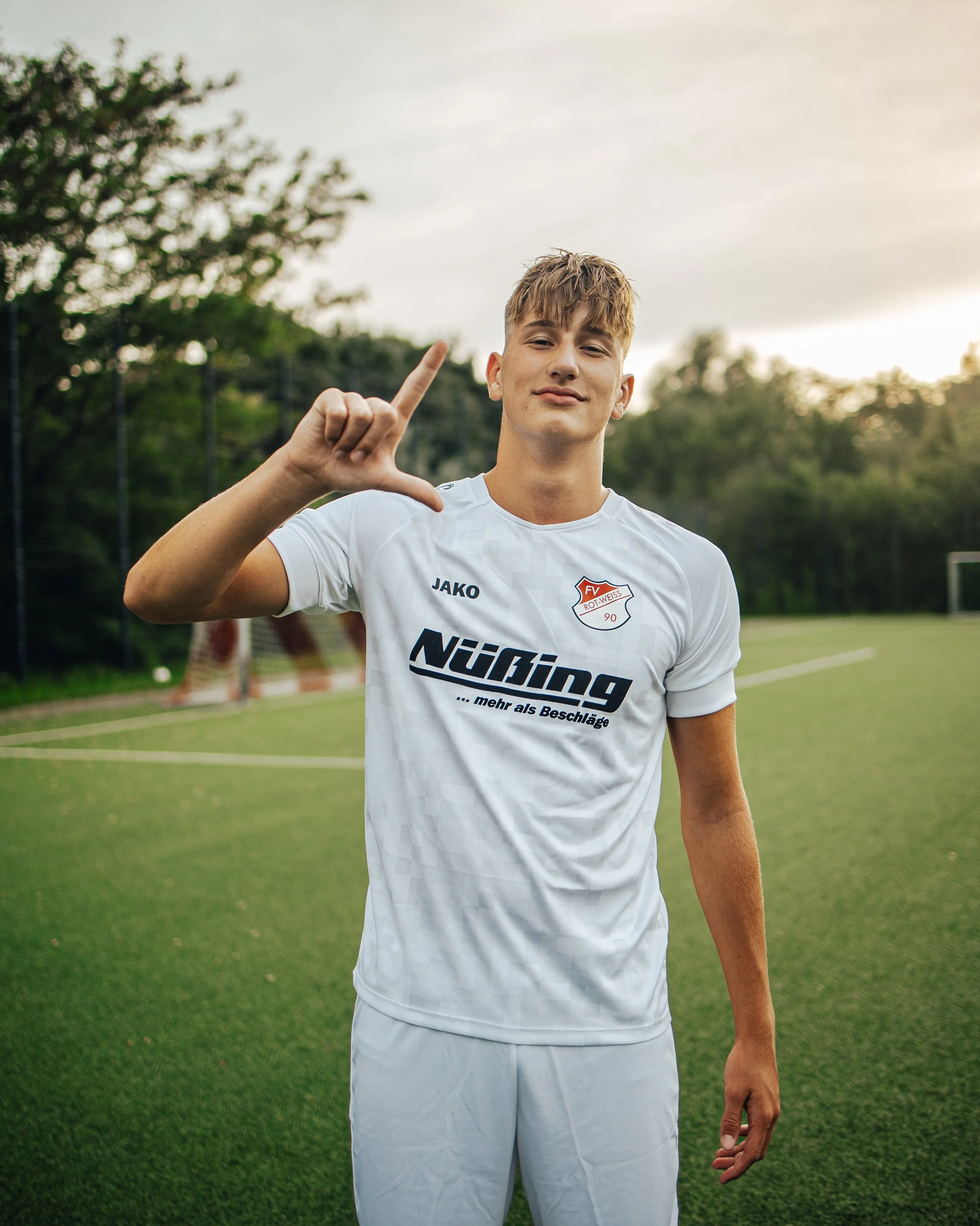 A young man in a white soccer uniform standing on a soccer field, making an L shape with his right hand, with trees and a goalpost in the background during sunset.
