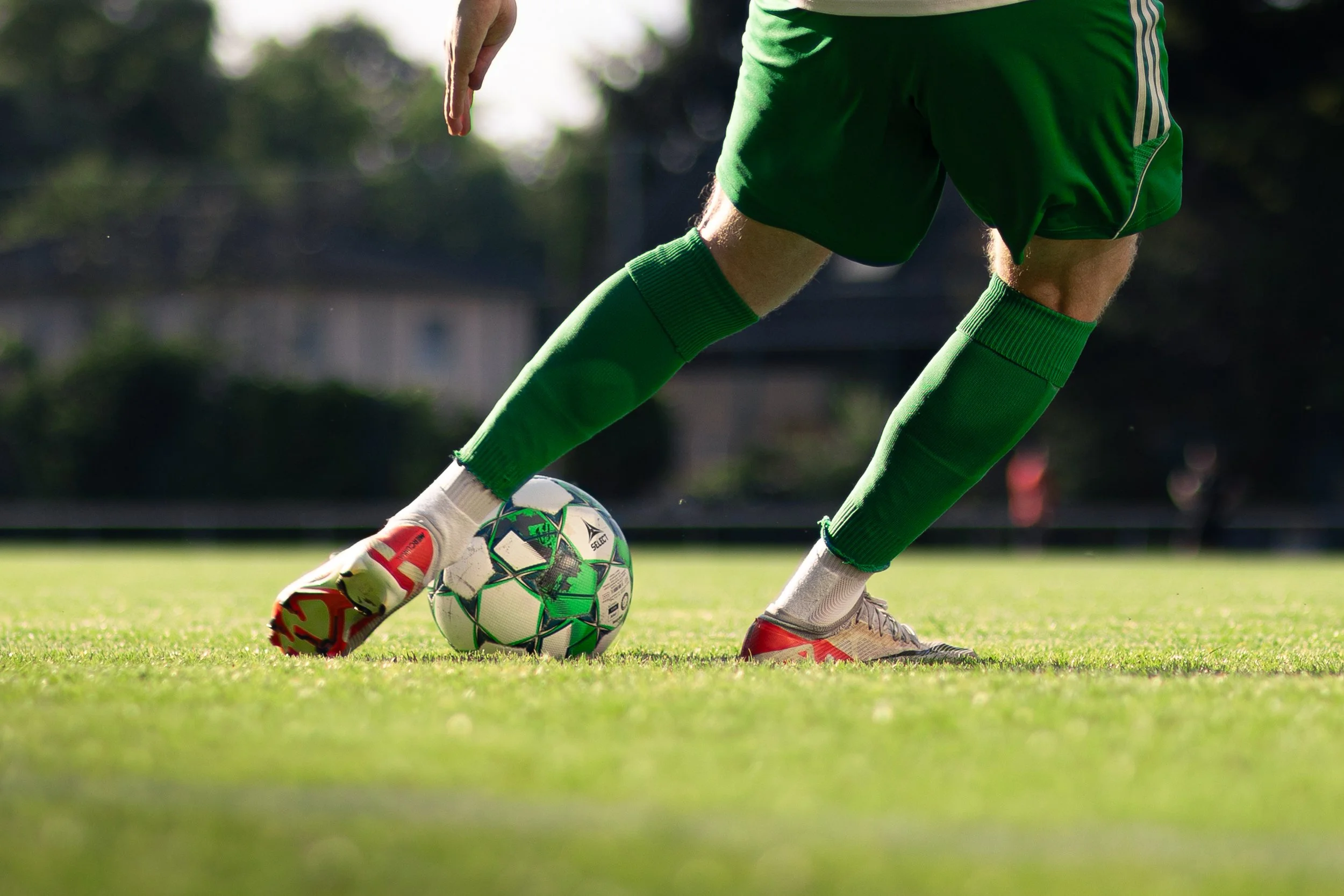 A soccer player in a green uniform and red cleats is kicking a soccer ball on a grassy field.
