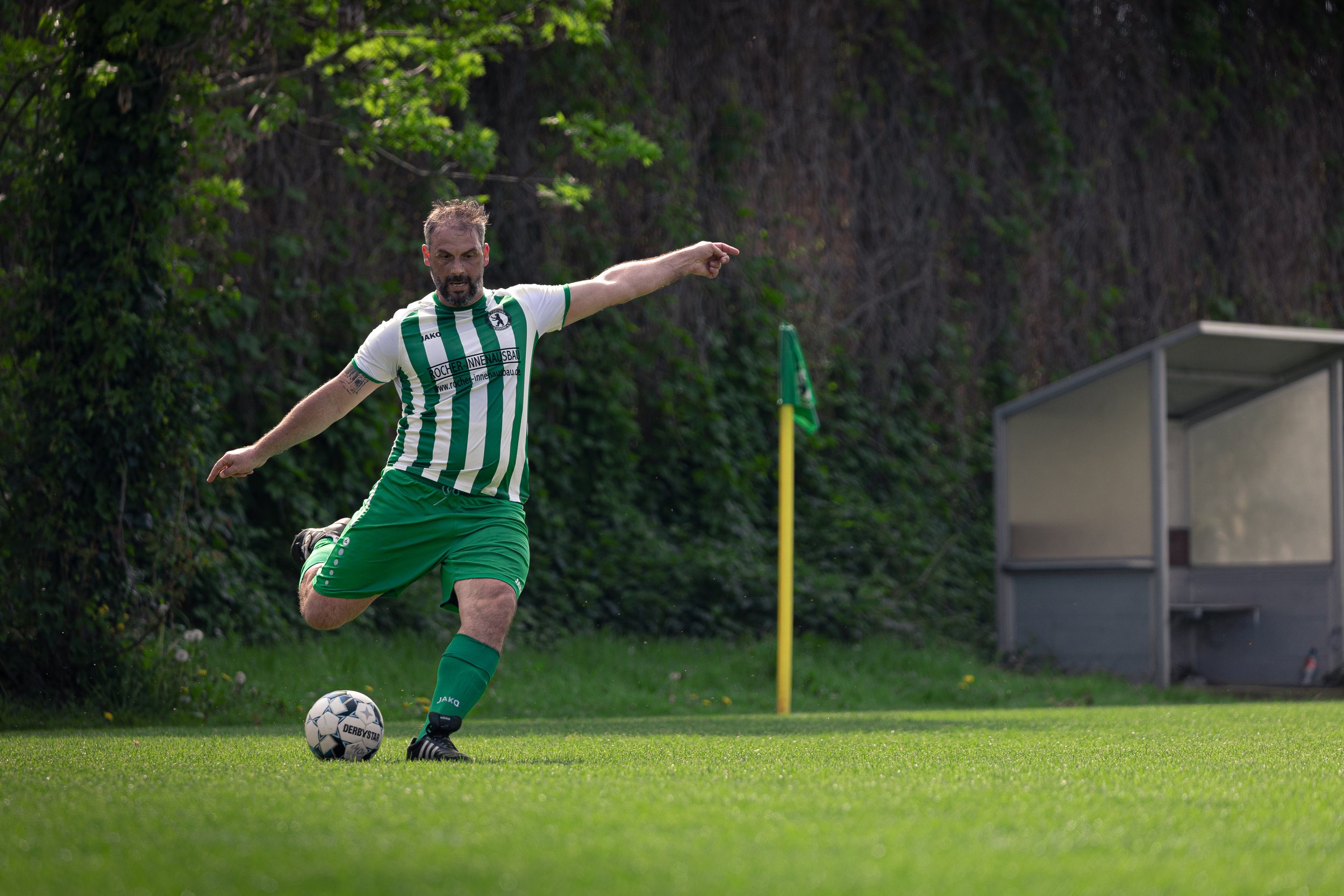 A man in a green and white striped soccer uniform kicking a soccer ball on a grassy field near a corner flag.