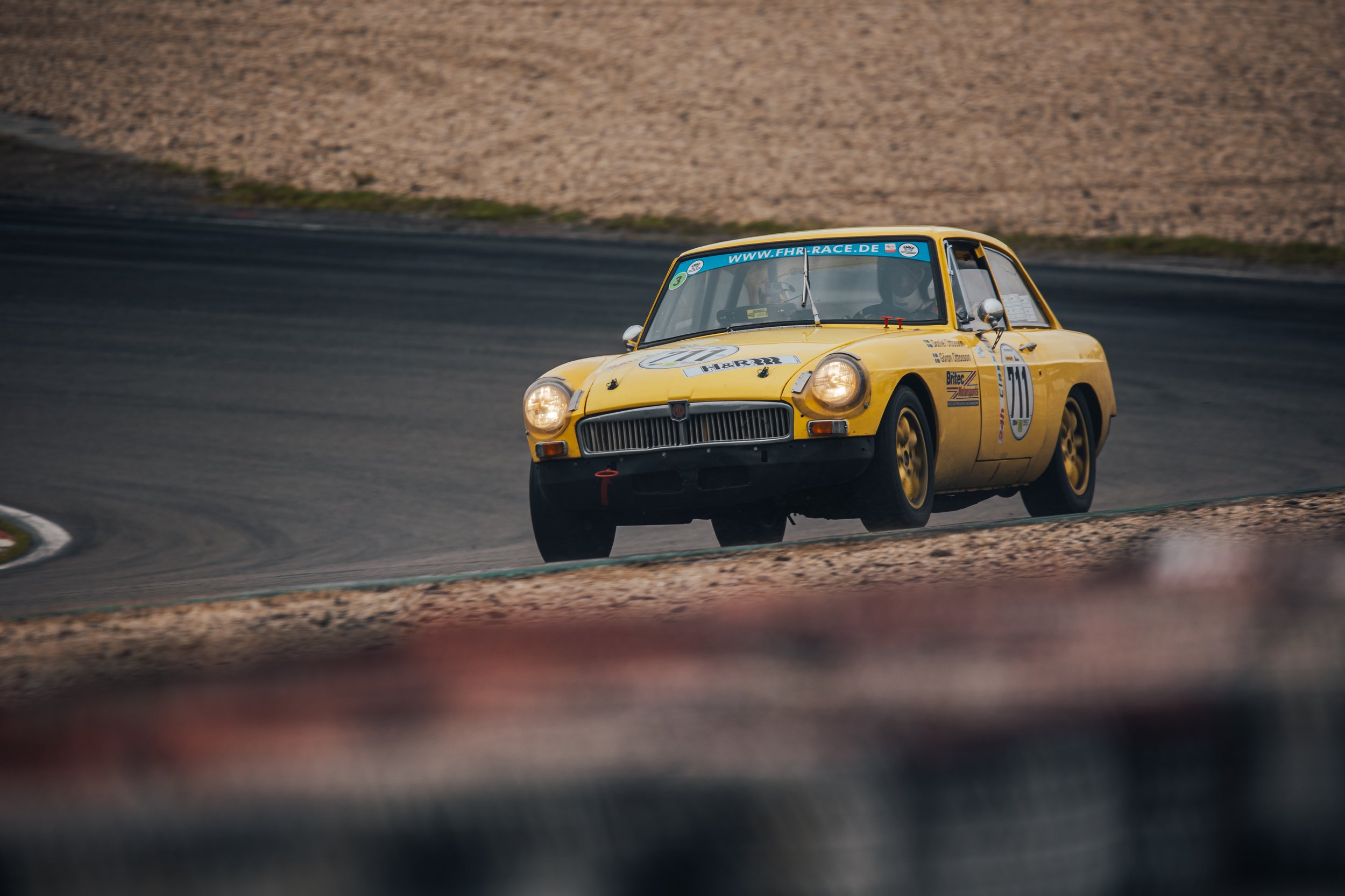 A yellow vintage race car on a racetrack, navigating a turn, with a gravelly area in the background.
