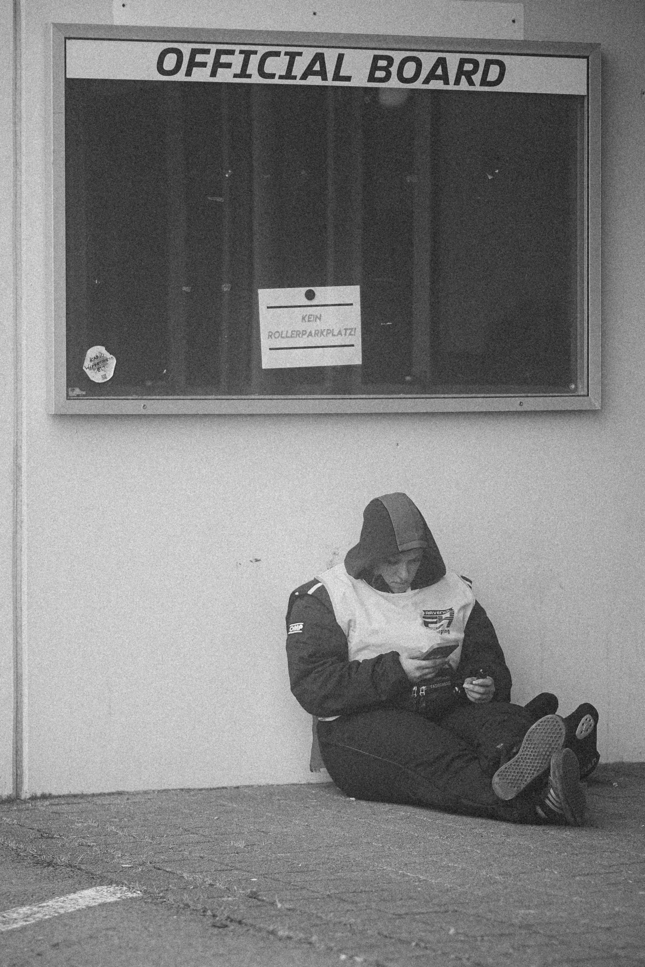 A person sitting on the ground against a wall, wearing a hoodie and jacket, looking at their phone. Above them is a sign that reads 'OFFICIAL BOARD' and a smaller note that says 'KEN ROLLER PARK PLAZA'.