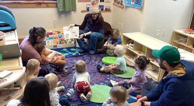A woman reading a book to a group of children in a classroom, with toys and shelves in the background.