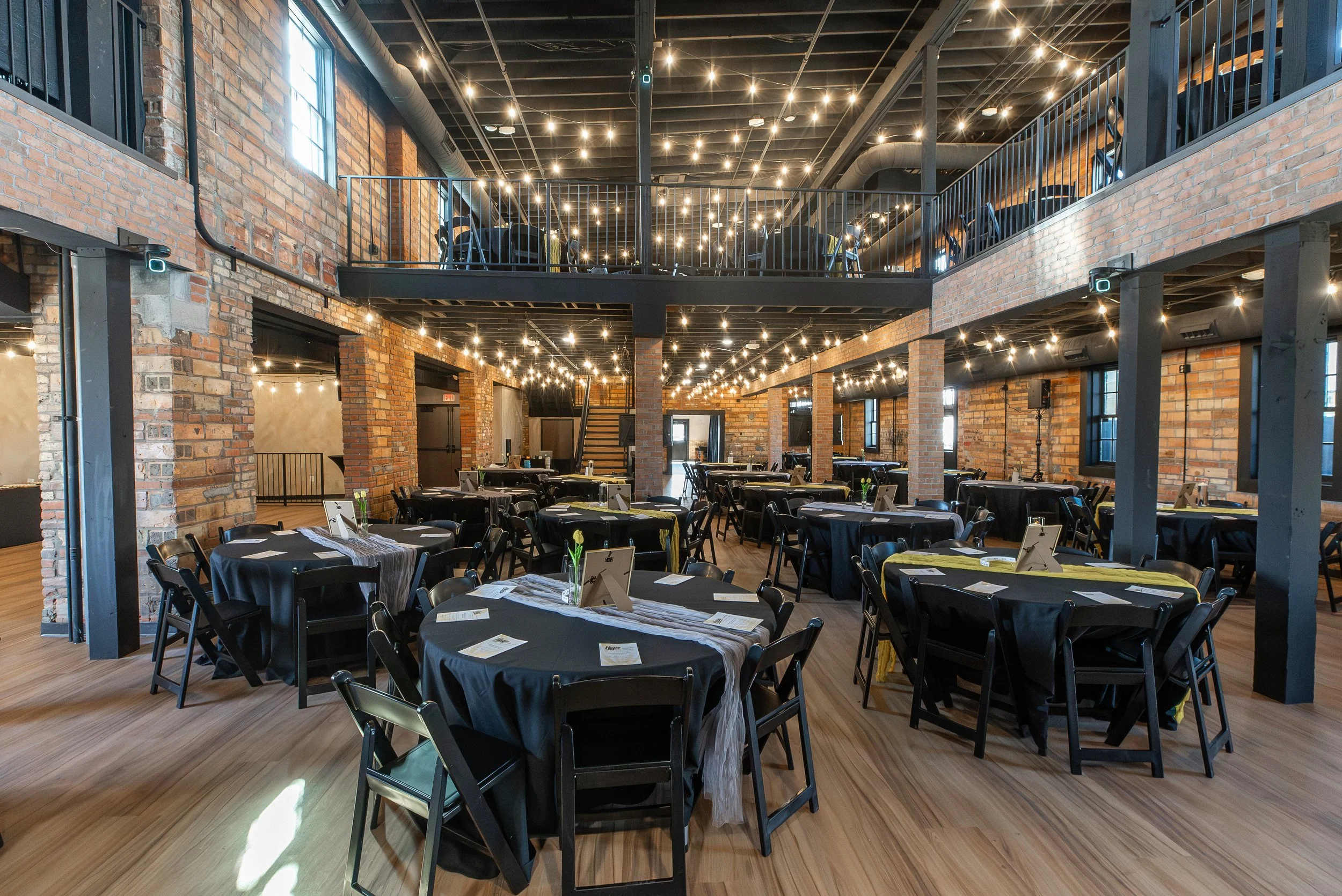 Interior of a modern event space with brick walls, black tables with black chairs, and string lights hanging from the ceiling.