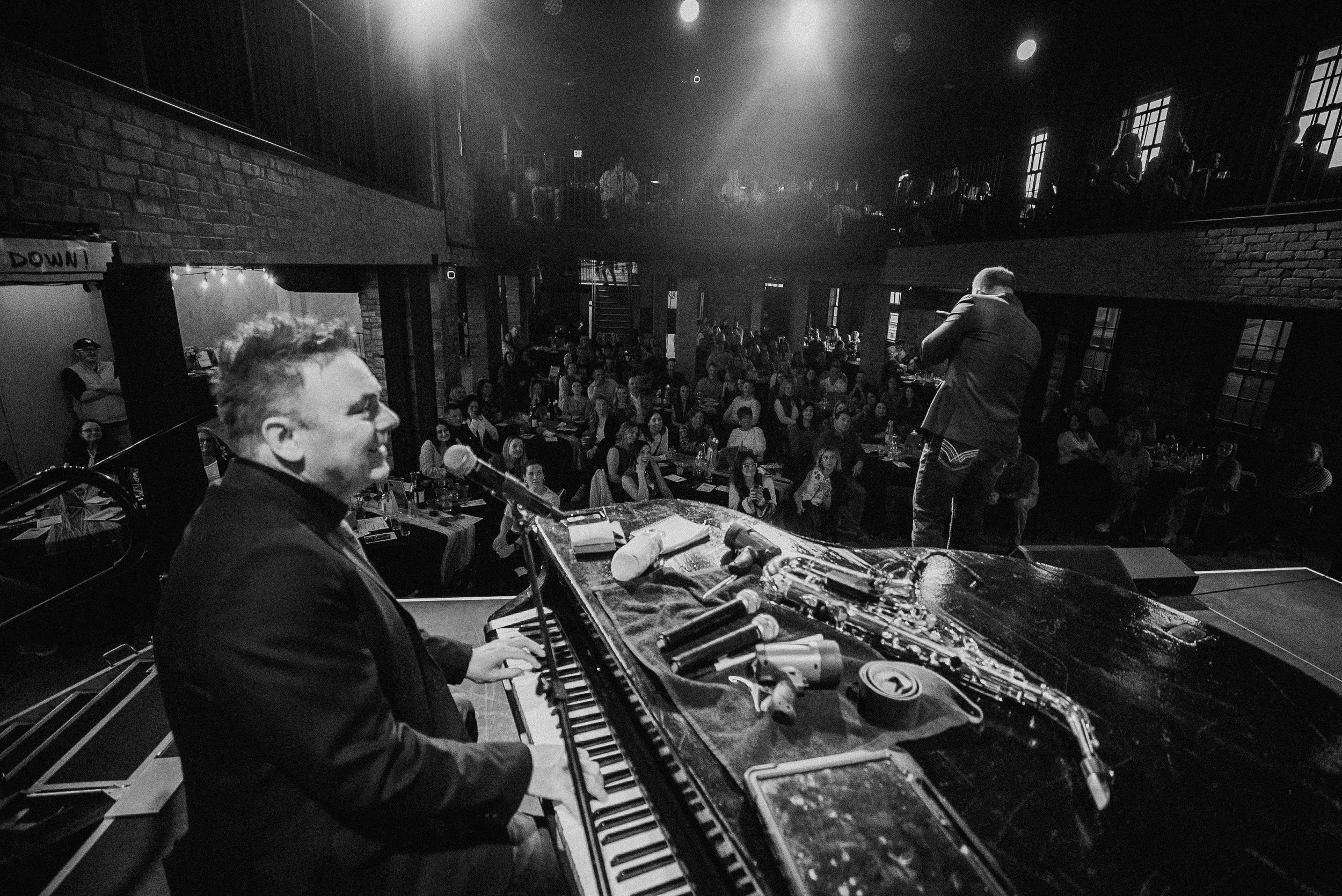 A jazz band performing live on stage with a pianist and a saxophonist, in front of an audience in a dark, intimate venue with brick walls and large windows.
