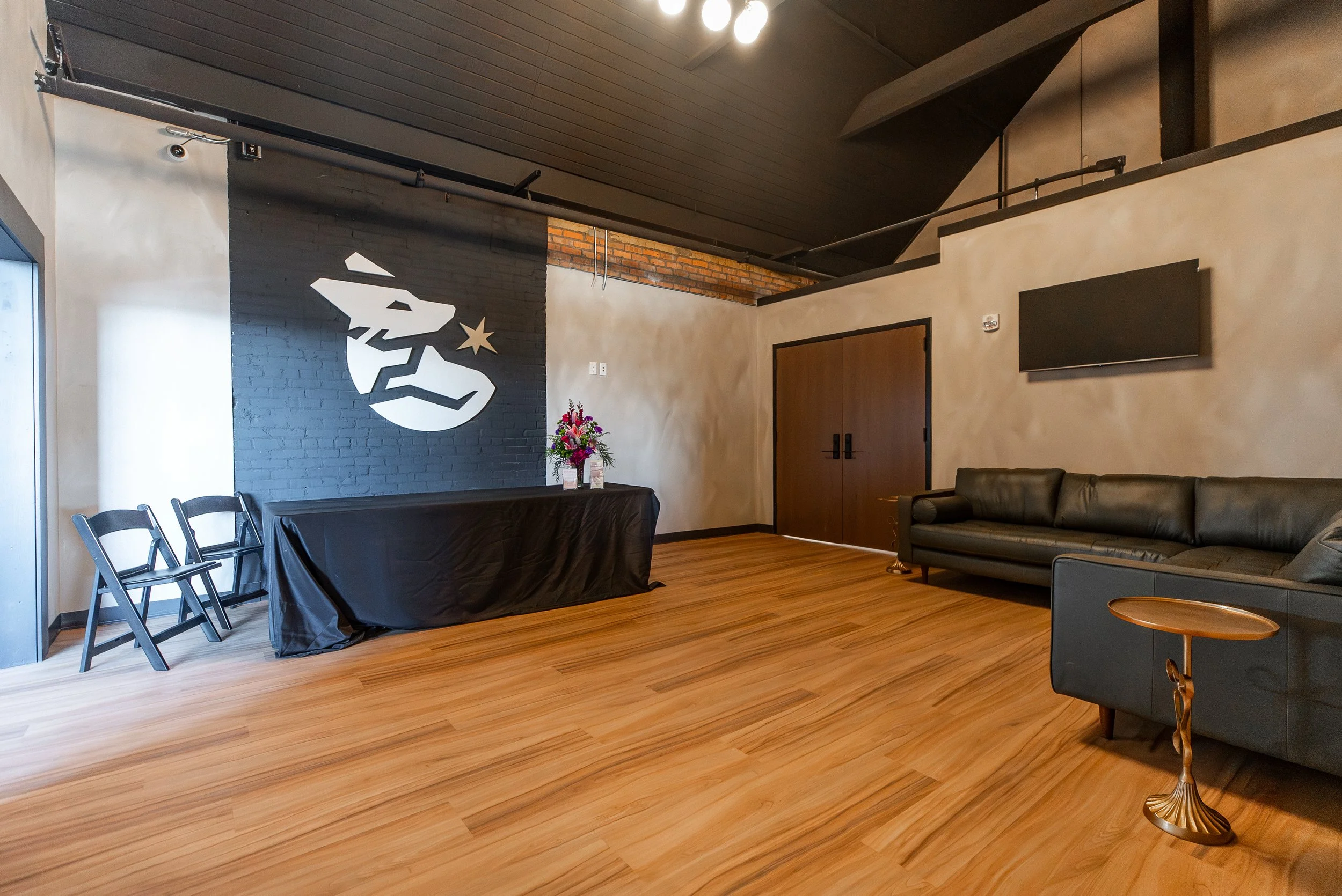 An indoor room with wood flooring, a black wall with a white face and star mural, a black table with floral arrangement, black chairs, a gray sofa, a flat-screen TV, wooden double doors, and a sloped ceiling with black beams and ceiling lights.