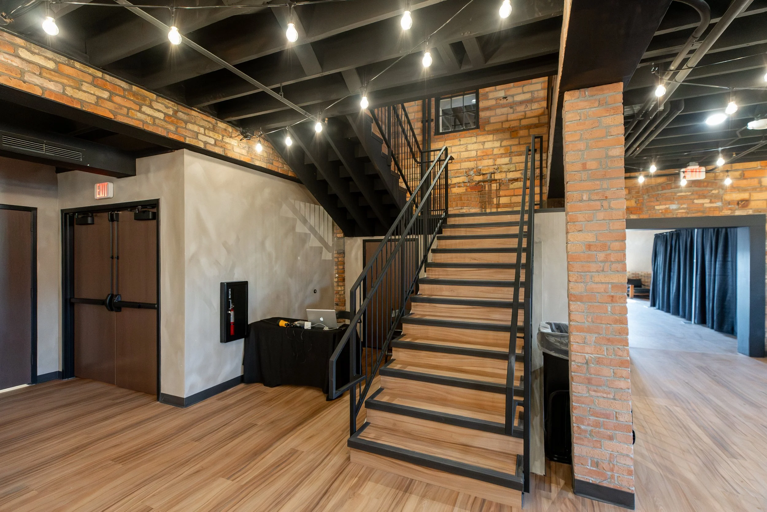 Interior of a modern industrial-style building with exposed brick walls, a staircase with wooden steps and black metal railing, string lights hanging from the ceiling, and hardwood flooring.
